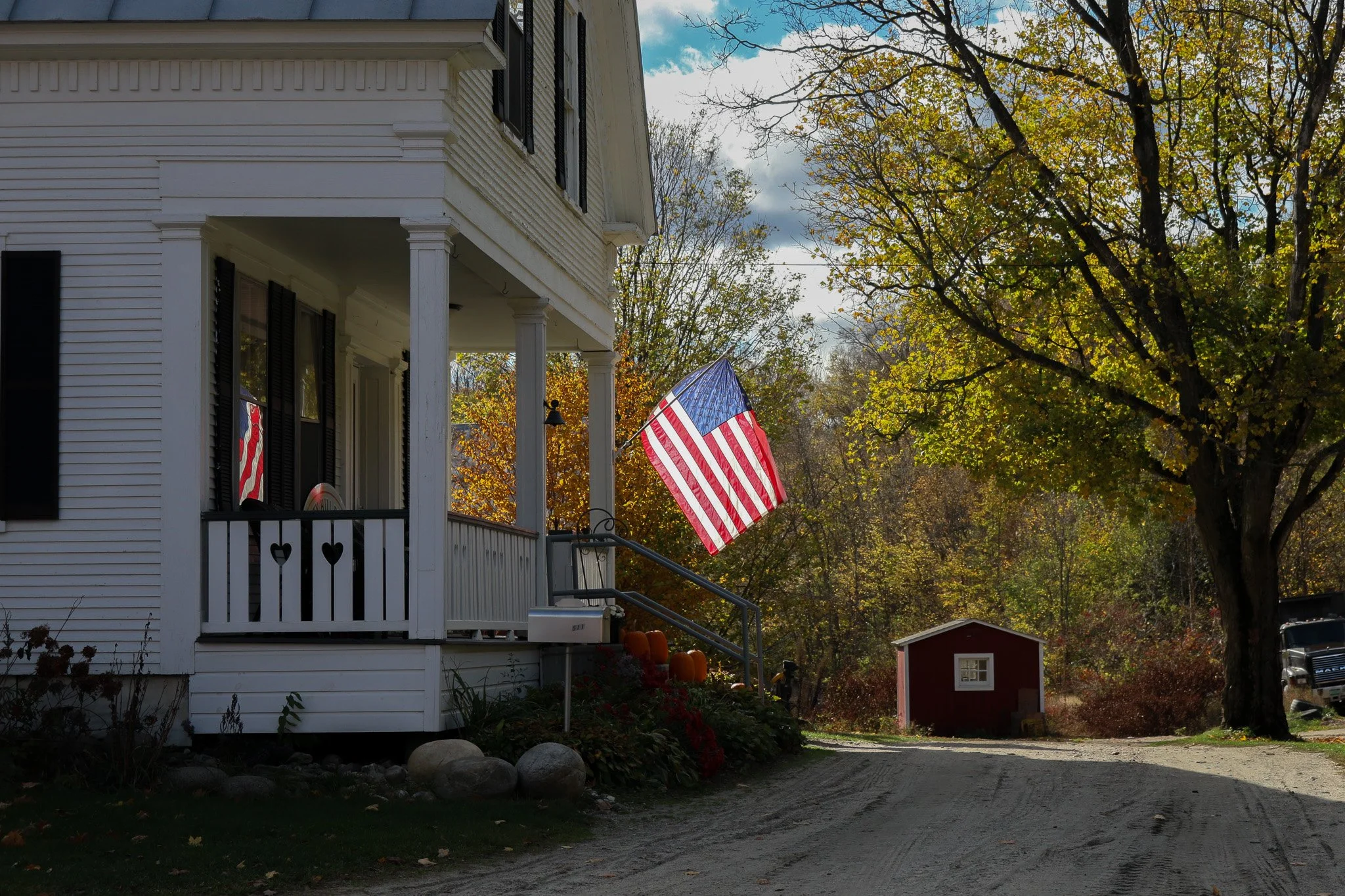 A white house with black shutters and a porch, decorated with pumpkins and a heart-shaped decor, has an American flag hanging outside. The scene is set in autumn with colorful trees and a gravel driveway leading to a small red shed.