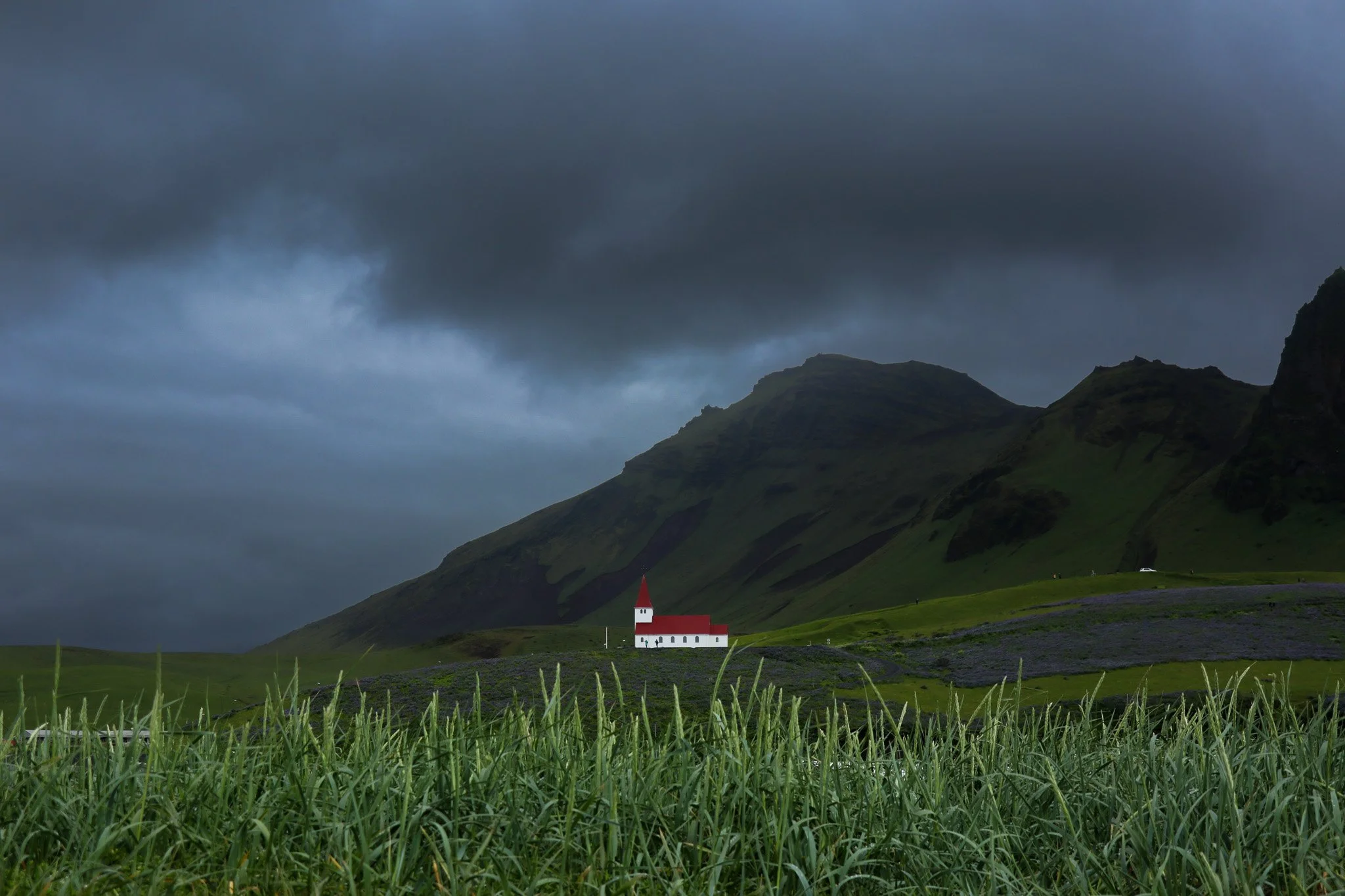 A small white church with a red roof and steeple situated in a green valley with tall grass in the foreground, dark mountains and overcast sky in the background.