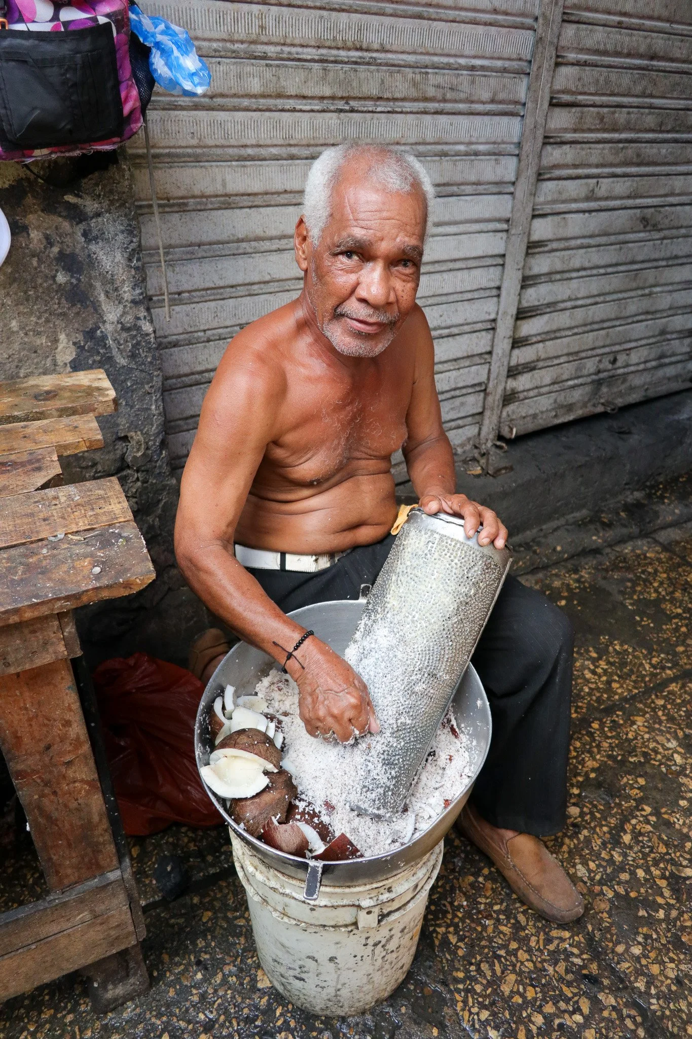 An elderly man with gray hair and a beard, shirtless, sitting on a chair, using a grater to grate coconut into a large bowl. The setting appears to be outdoors with a metal shutter in the background, and the ground is wet. The man is focused on his t