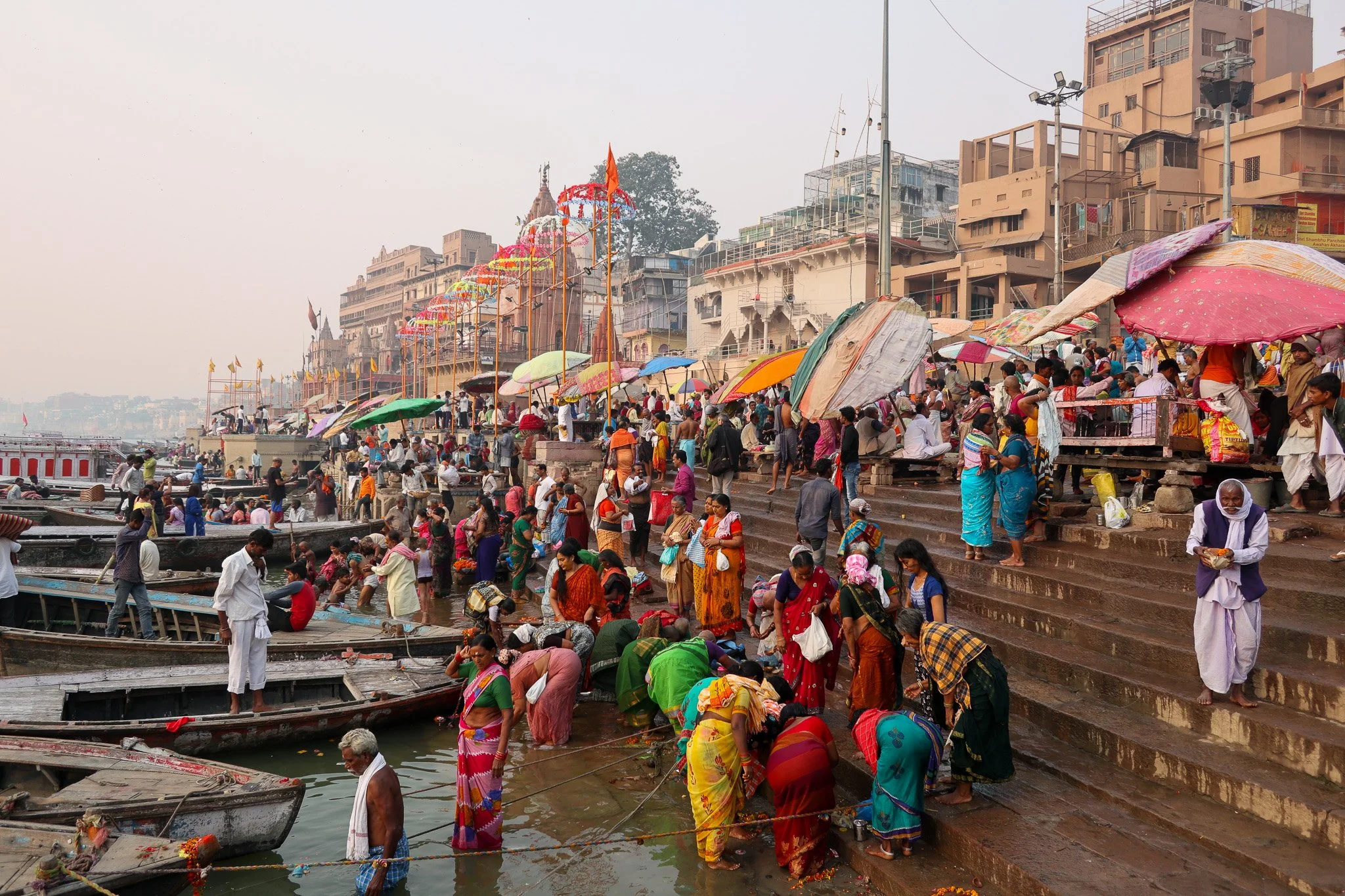 People gather along the steps and shoreline of a river, with boats docked nearby. Many individuals are wearing colorful traditional clothing and carrying umbrellas. The scene includes a busy crowd engaging in a ritual or ceremony, set against buildin