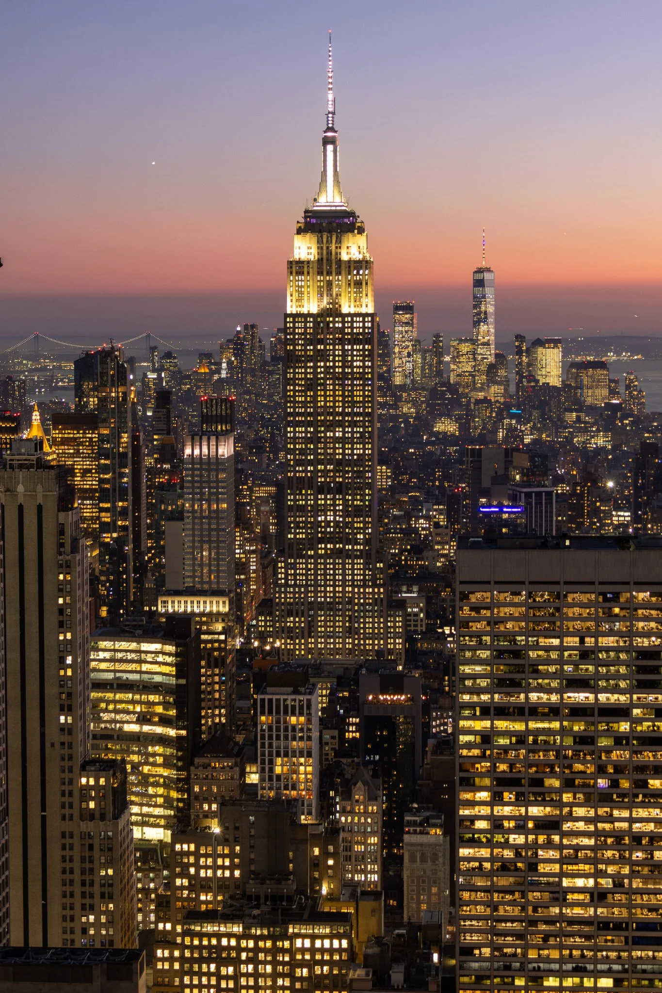 Nighttime cityscape of New York City with the Empire State Building illuminated prominently in the center, and One World Trade Center visible in the distance to the right.