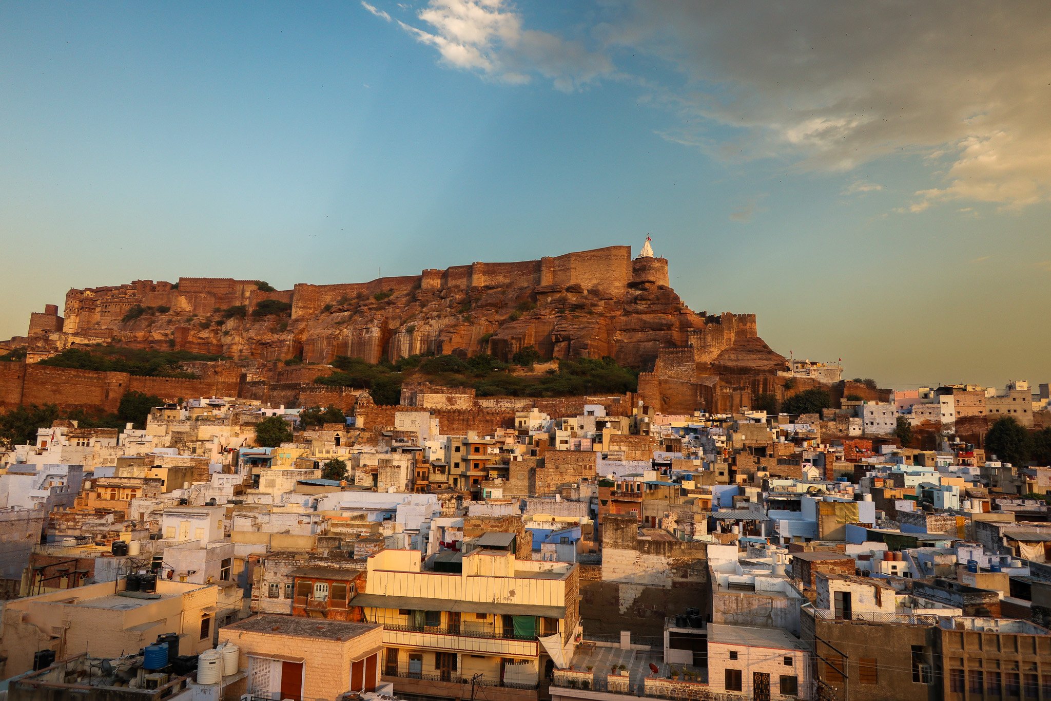 A cityscape with tightly packed buildings and a large hill with fortress walls and a white monument on top, under a partly cloudy sky.