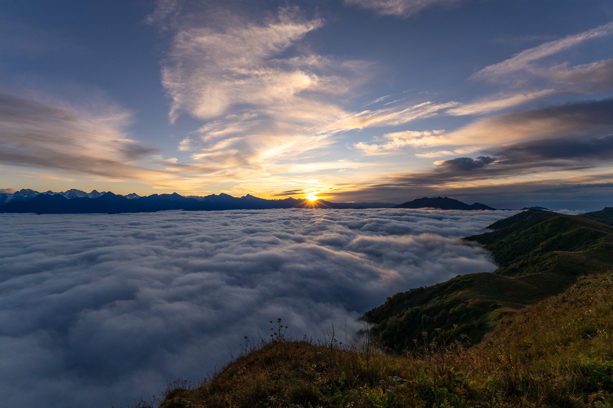 Sunrise over a mountain range with clouds covering the valleys and green hillside in the foreground.