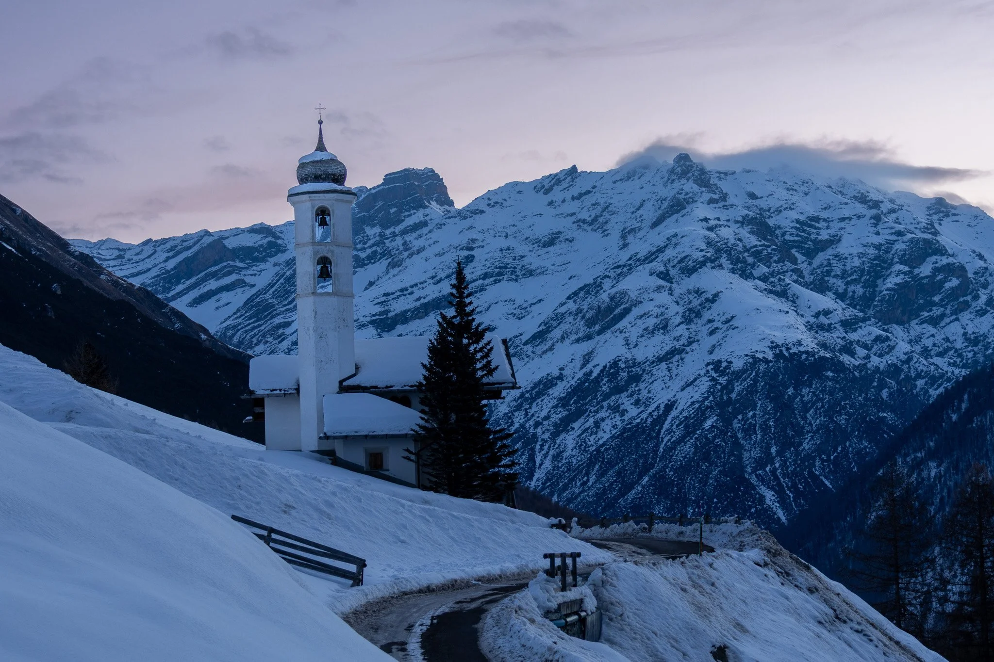 A snow-covered mountain landscape with a white church featuring a bell tower and a golden dome, surrounded by snow and a partly cloudy sky.