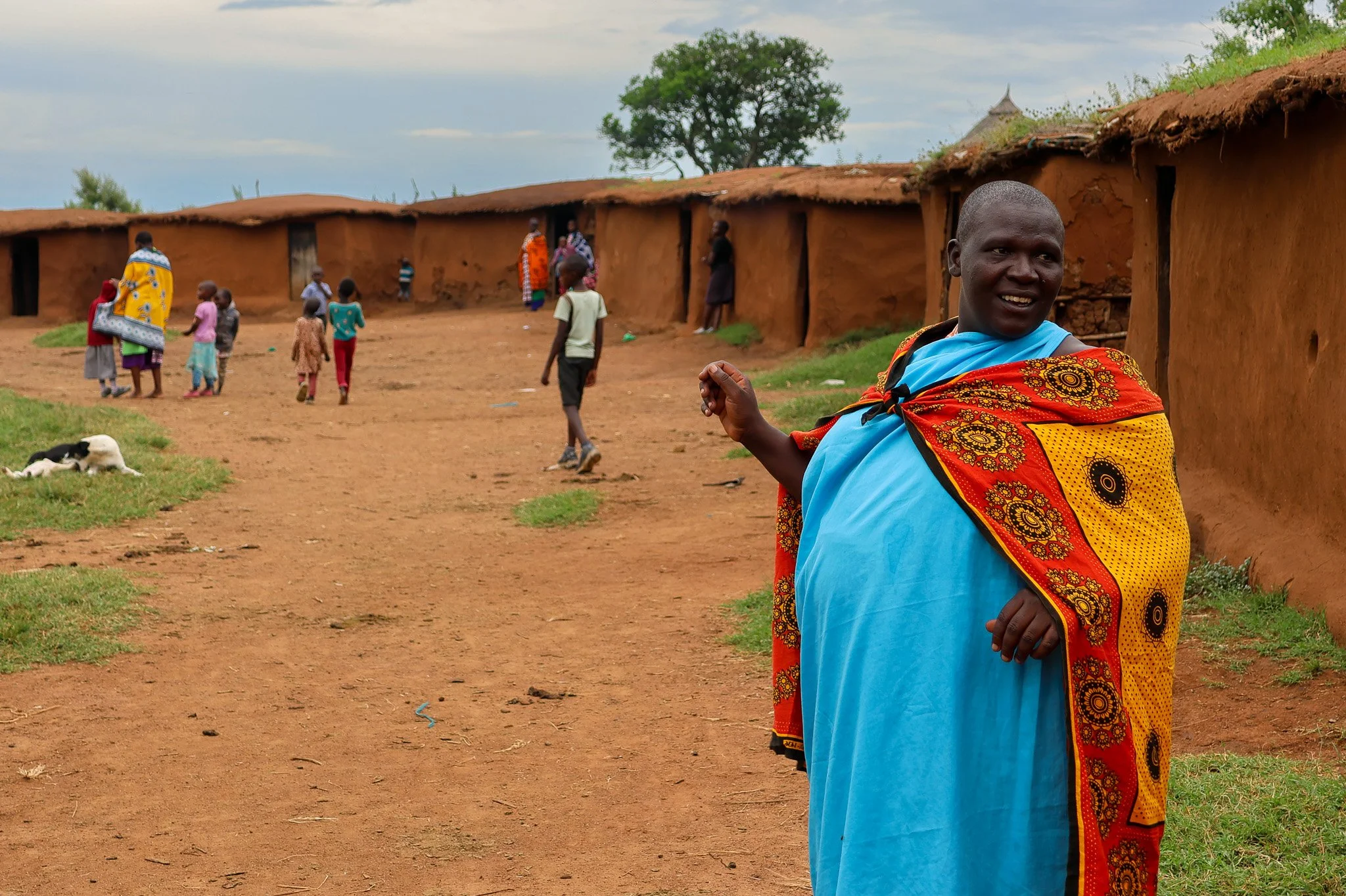 A woman in colorful traditional clothing standing outside a mud-brick village, with children and other adults in the background.