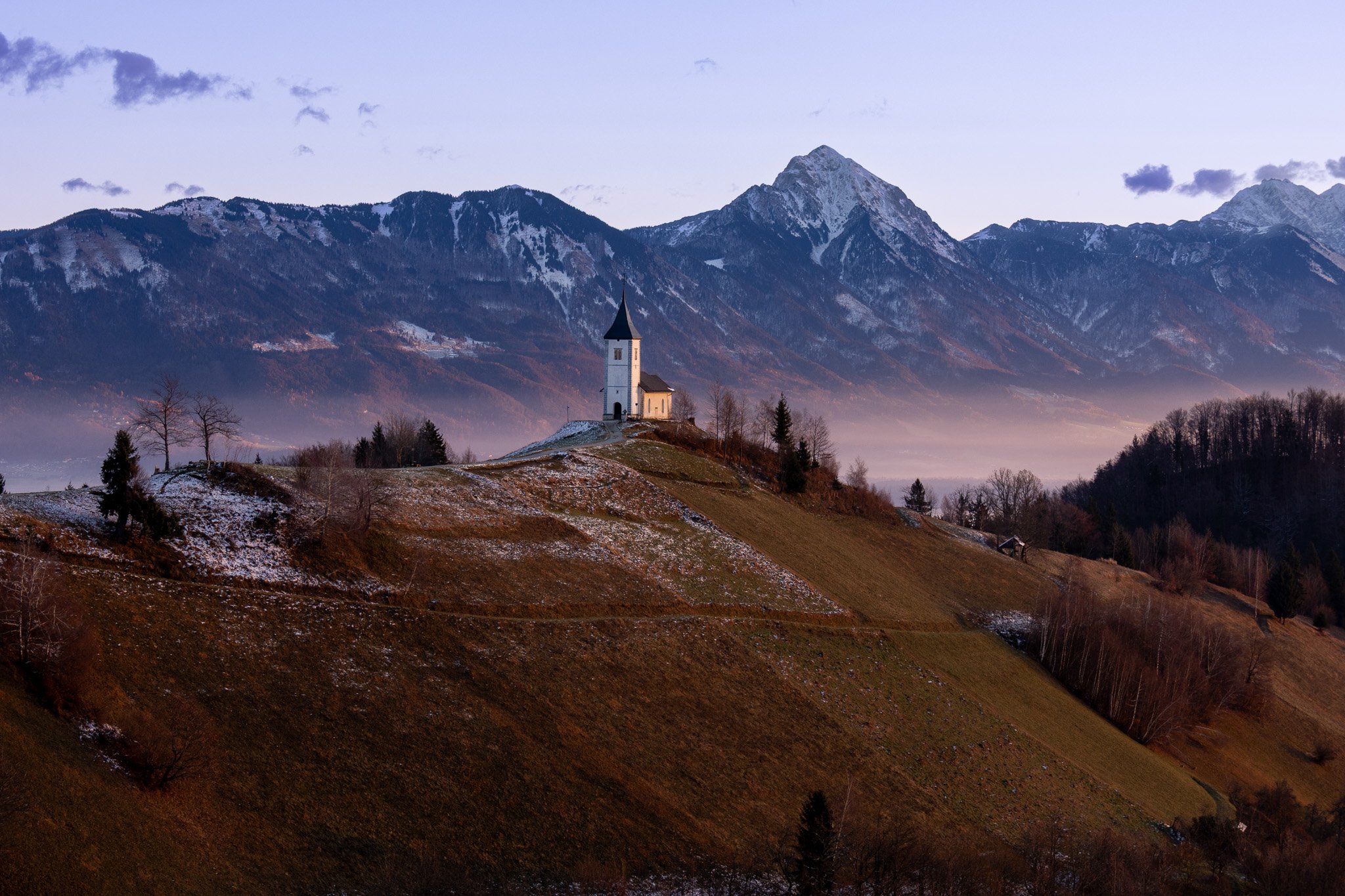 A small white church with a pointed black roof is situated on a hill with some patches of snow, overlooking a mountainous landscape with snow-capped peaks in the background.