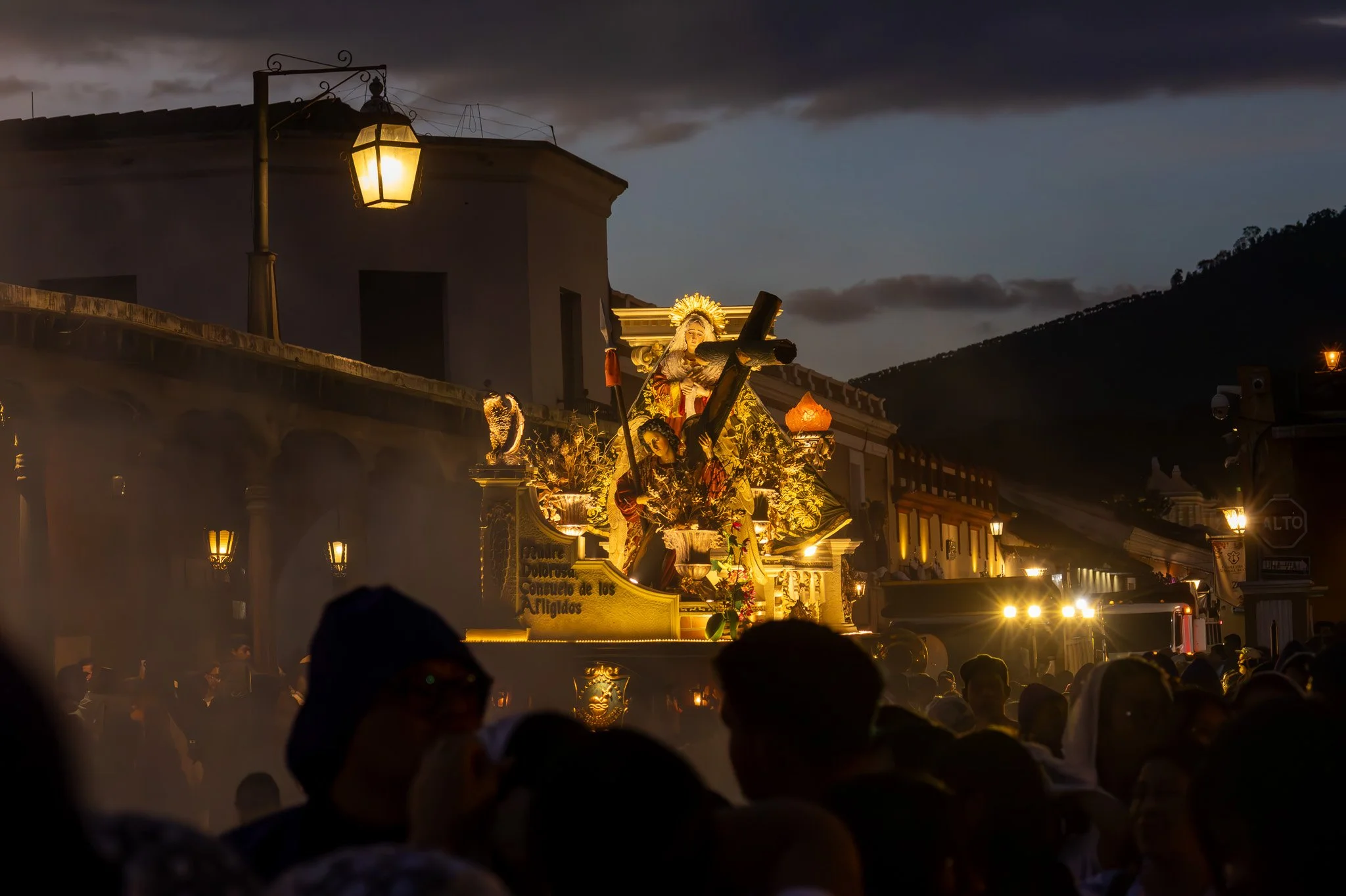 A religious procession during dusk with a float depicting Jesus Christ carrying a cross, surrounded by flowers and statues, in front of a crowd of people on a street illuminated by street lamps.
