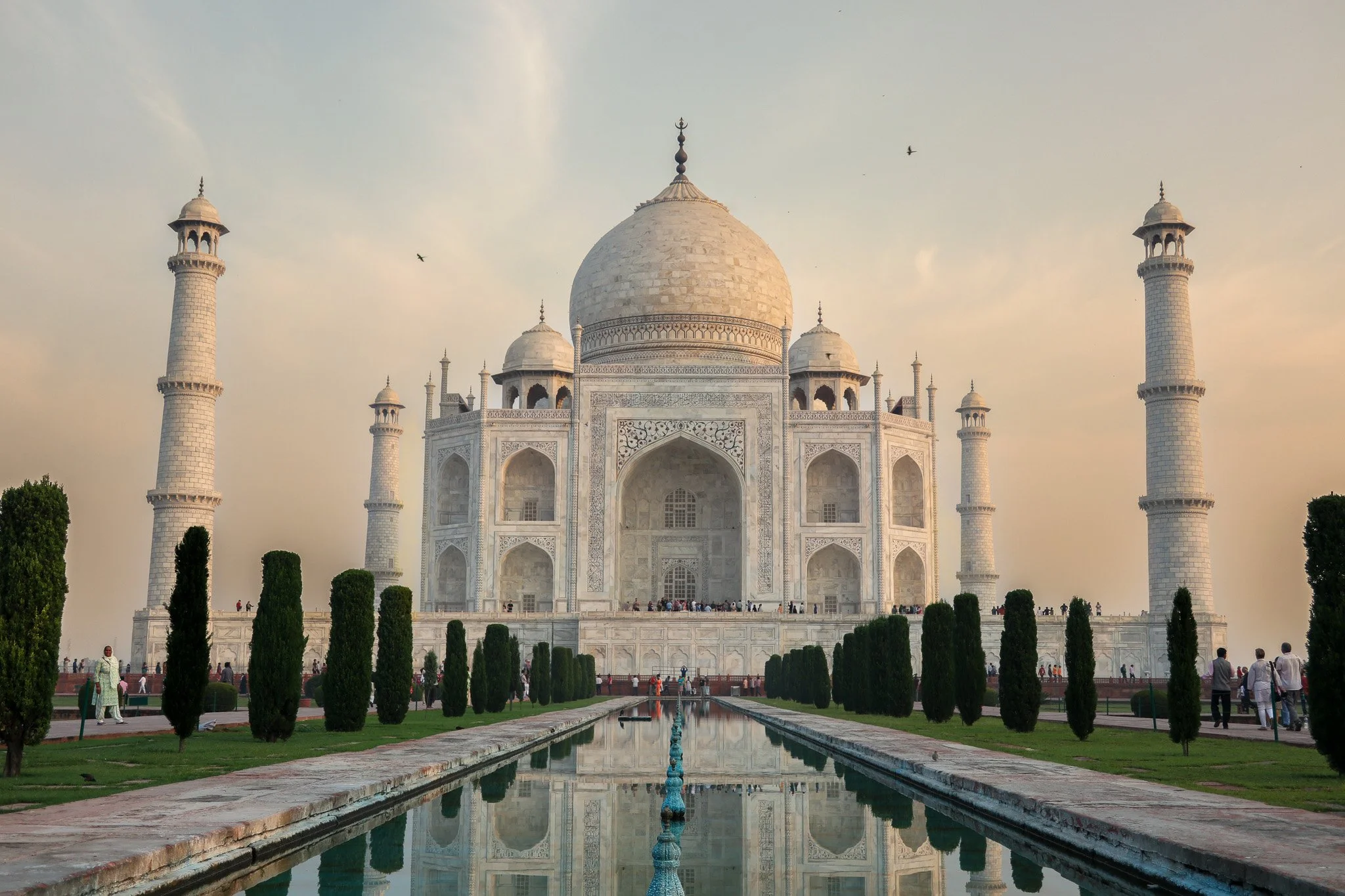 The Taj Mahal, a white marble mausoleum with a large central dome and four minarets, surrounded by a garden with a reflecting pool and visitors walking around.