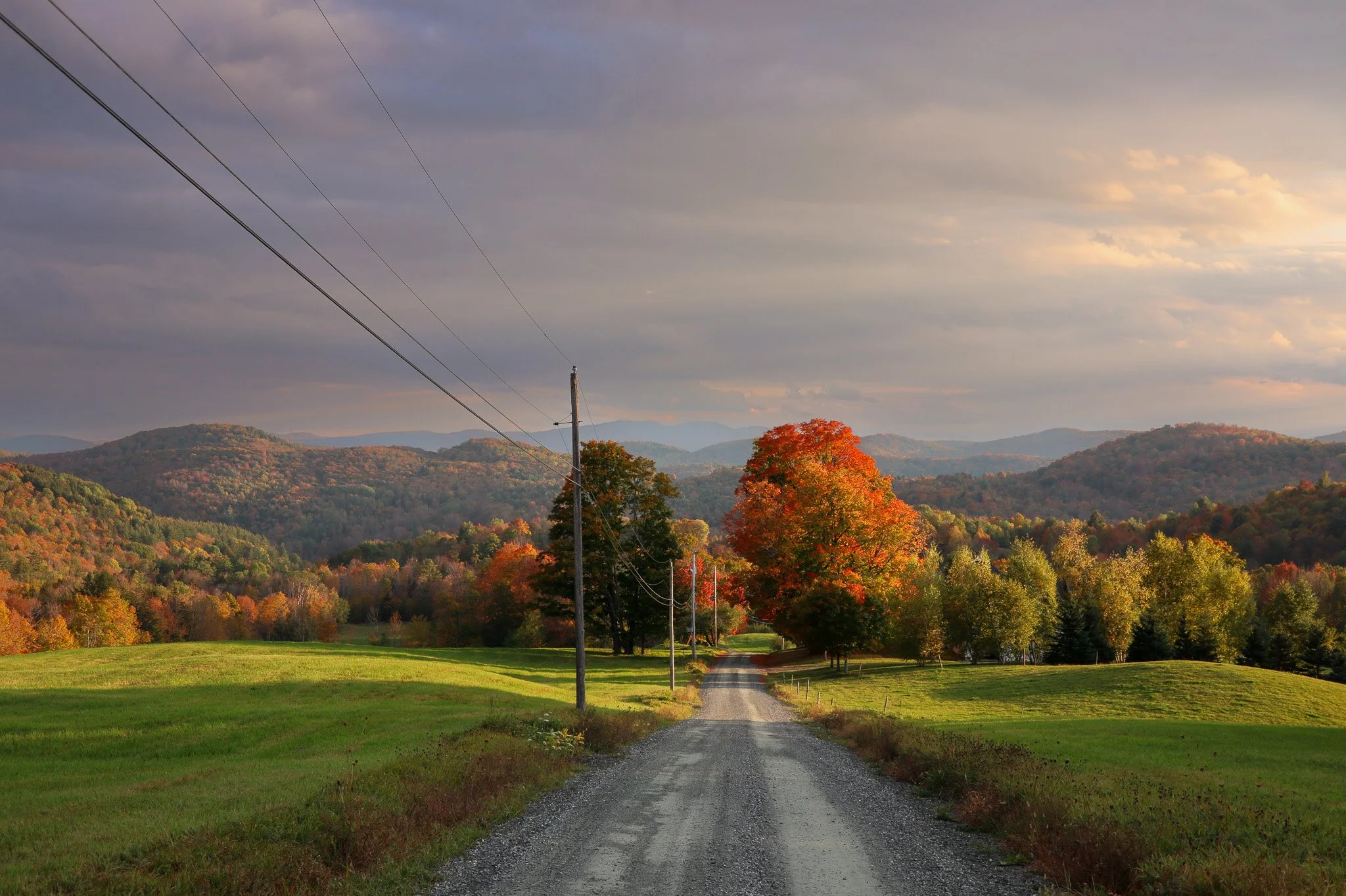 A gravel country road lined with power lines, leading towards colorful autumn trees and distant mountains in the background under a cloudy sky.