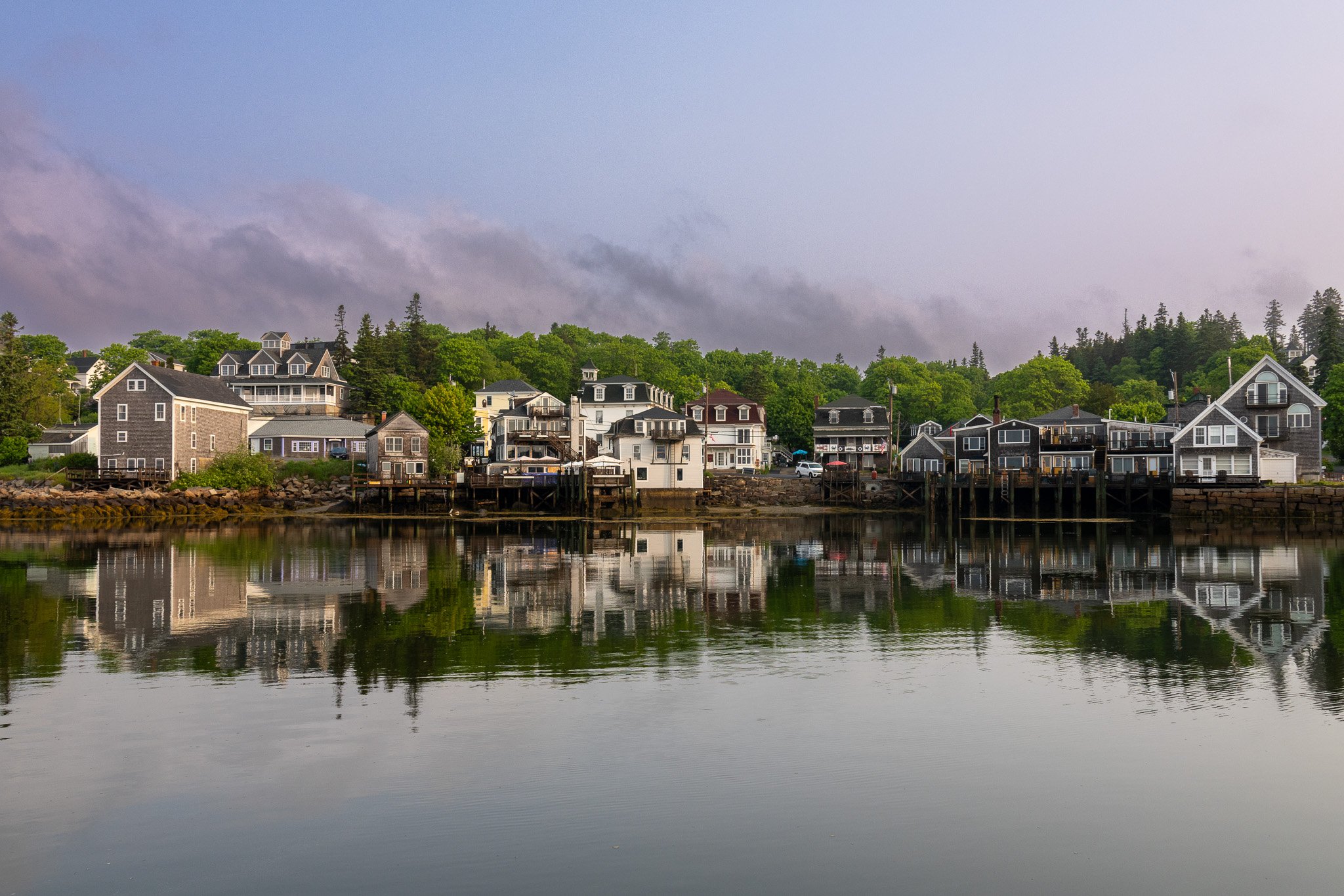 A row of houses along the water with their reflections visible in the calm water below, trees and hills in the background, and a cloudy sky overhead.