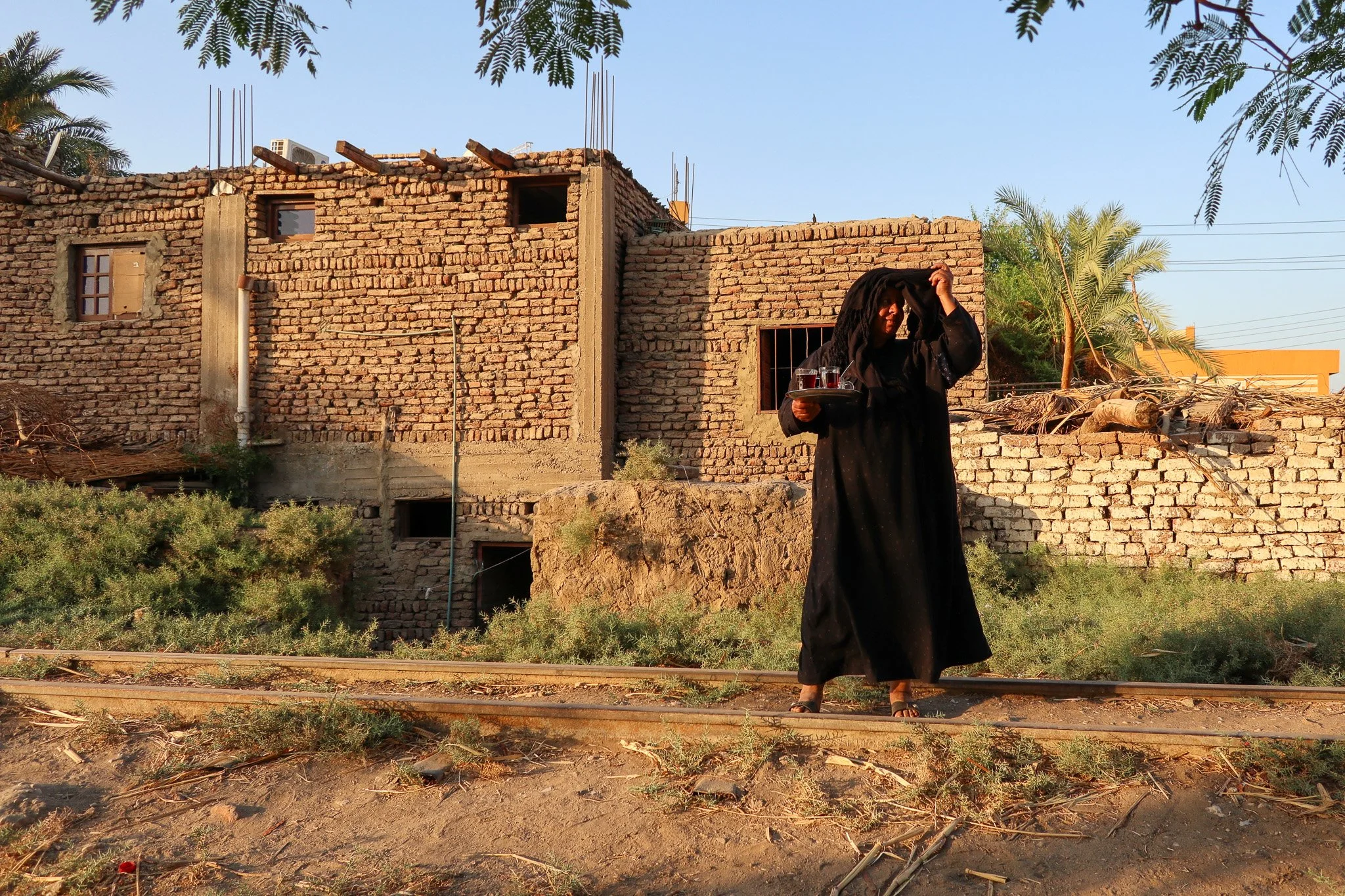 Woman in black traditional dress standing on railway track in front of unfinished brick building, holding tray with glasses of drinks, adjusting her headscarf, with sunlight and green trees around.
