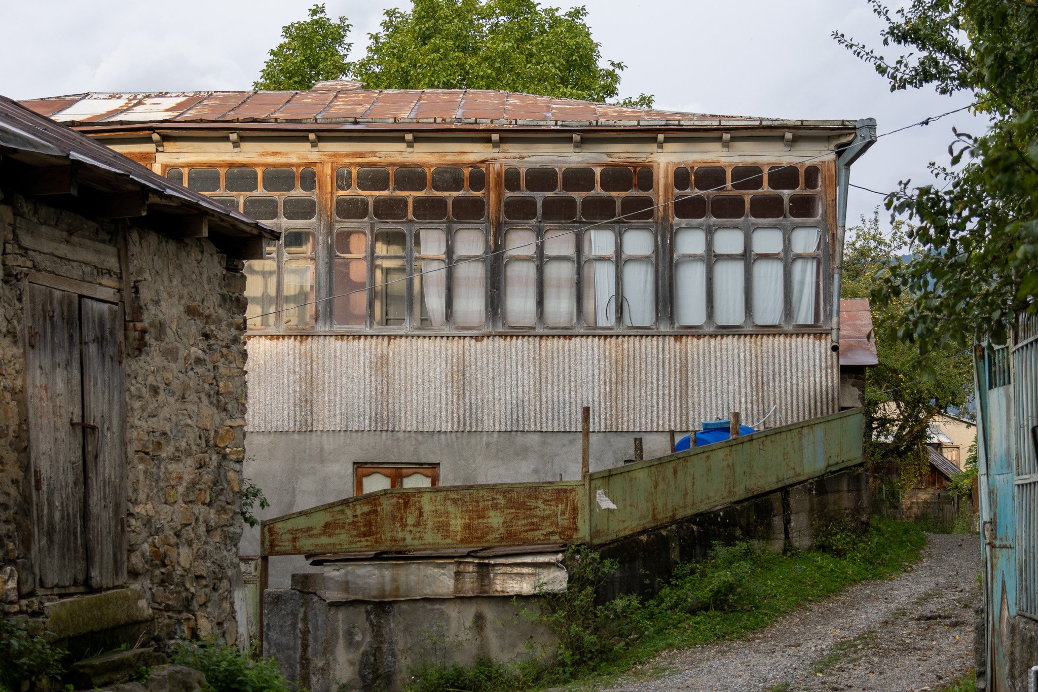 Old weathered house with a corrugated metal lower wall and a framed upper section with multiple small windows covered with white curtains. A rusty boat is placed on a stone foundation in front. Surroundings include stone and wooden structures, a grav