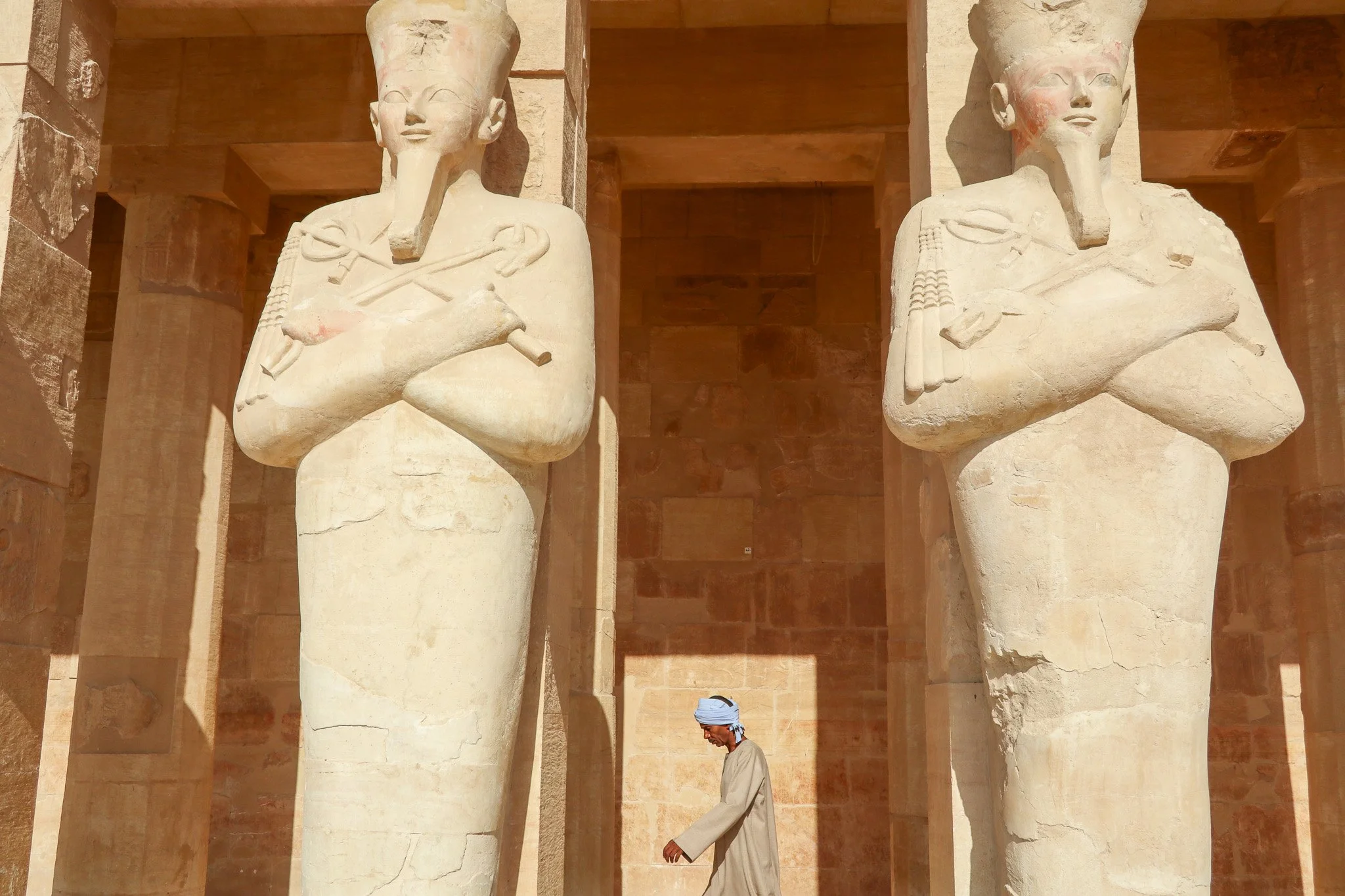 A person wearing traditional clothing and a head covering walking between two large ancient Egyptian statues with cat faces, holding crossed arms, in a stone temple