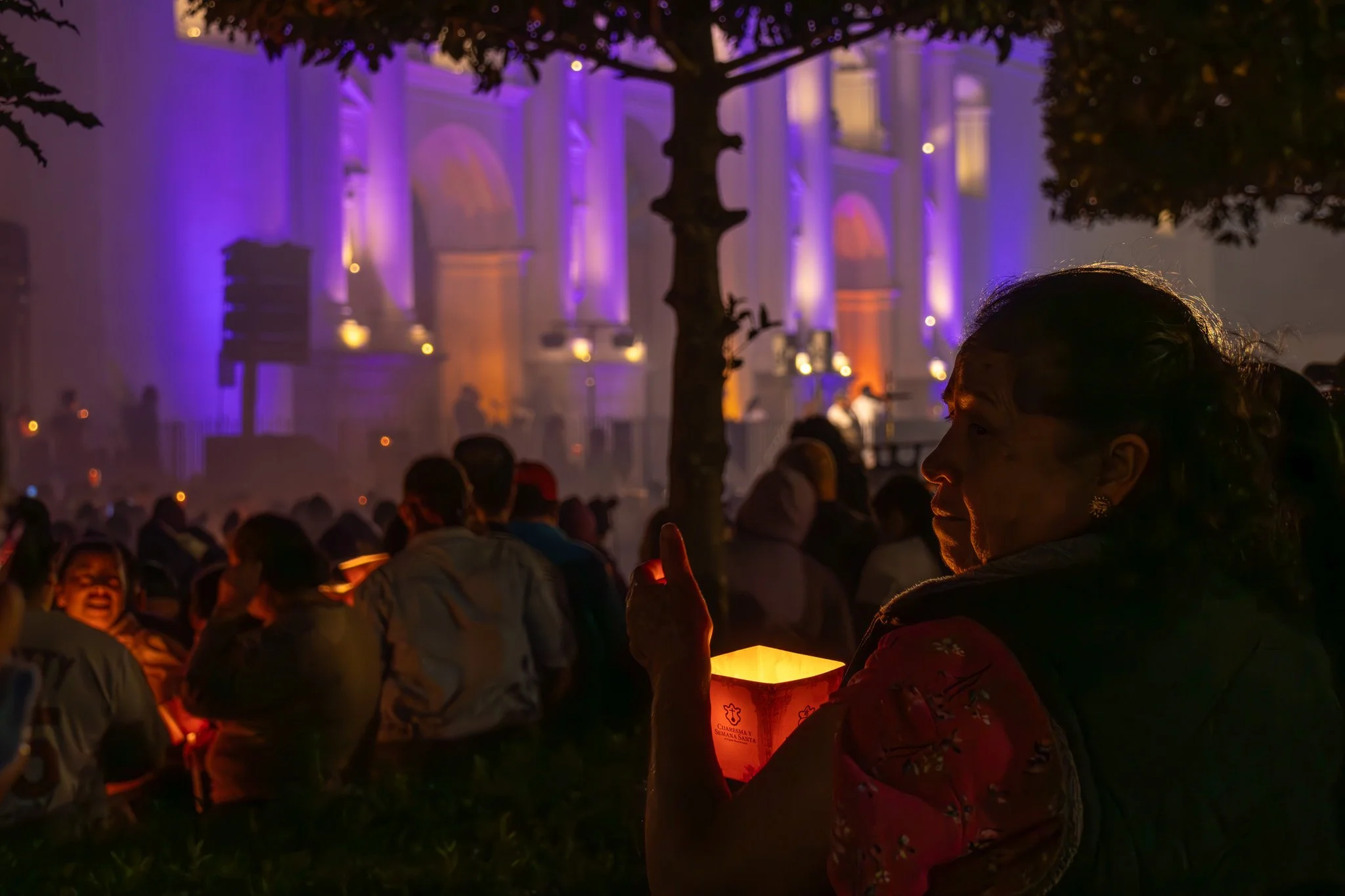 A woman holding a lit paper lantern at night during a large outdoor gathering, with a brightly lit, colorful building in the background.