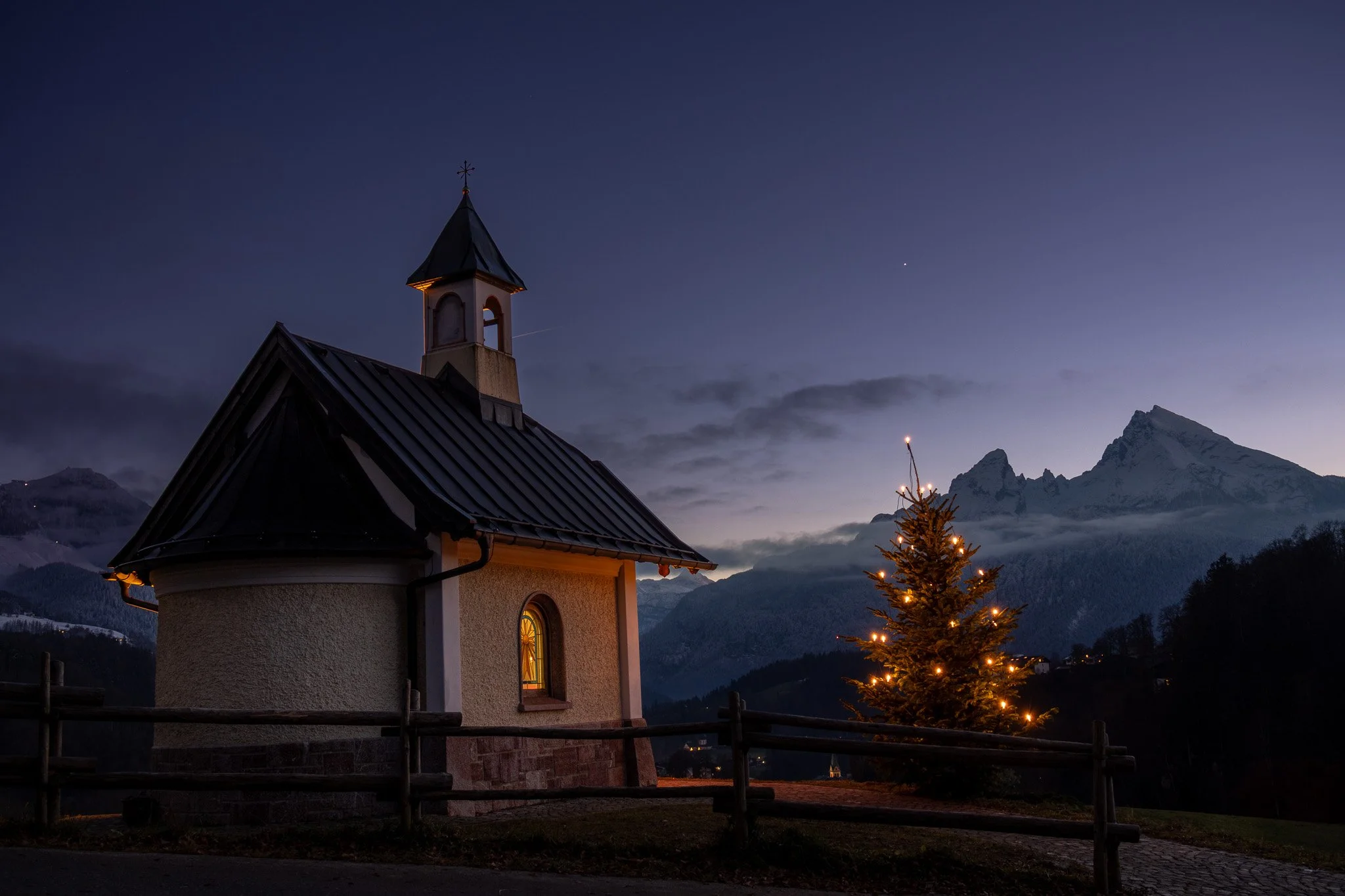 A small church with a steeple and a window illuminated from inside, surrounded by a wooden fence, with a decorated Christmas tree and snow-covered mountains in the background, during dusk.