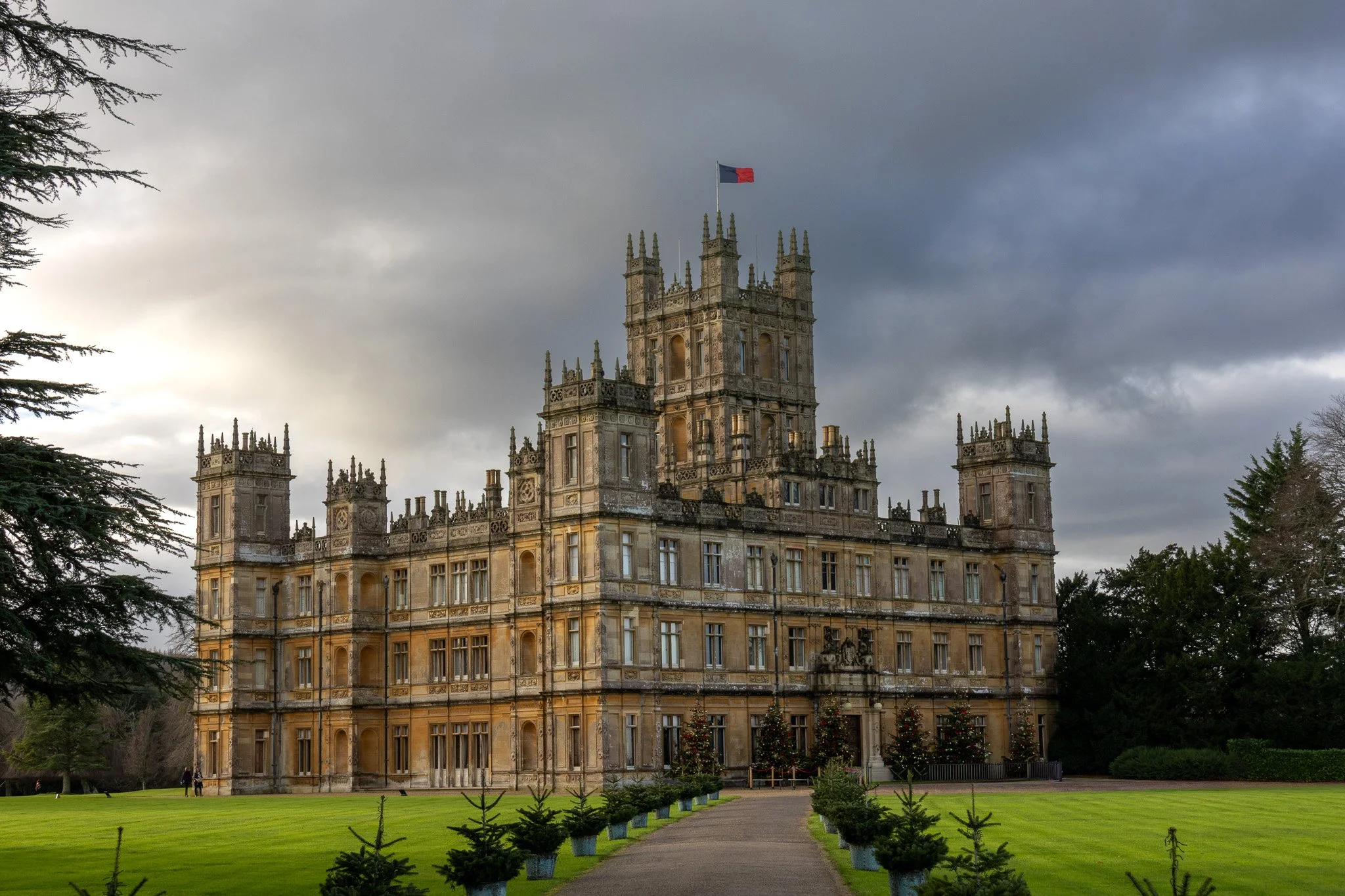 A large, historic castle with multiple turrets and a flag on top, situated in a well-manicured garden with a pathway leading to the entrance, trees on either side, and a cloudy sky overhead.