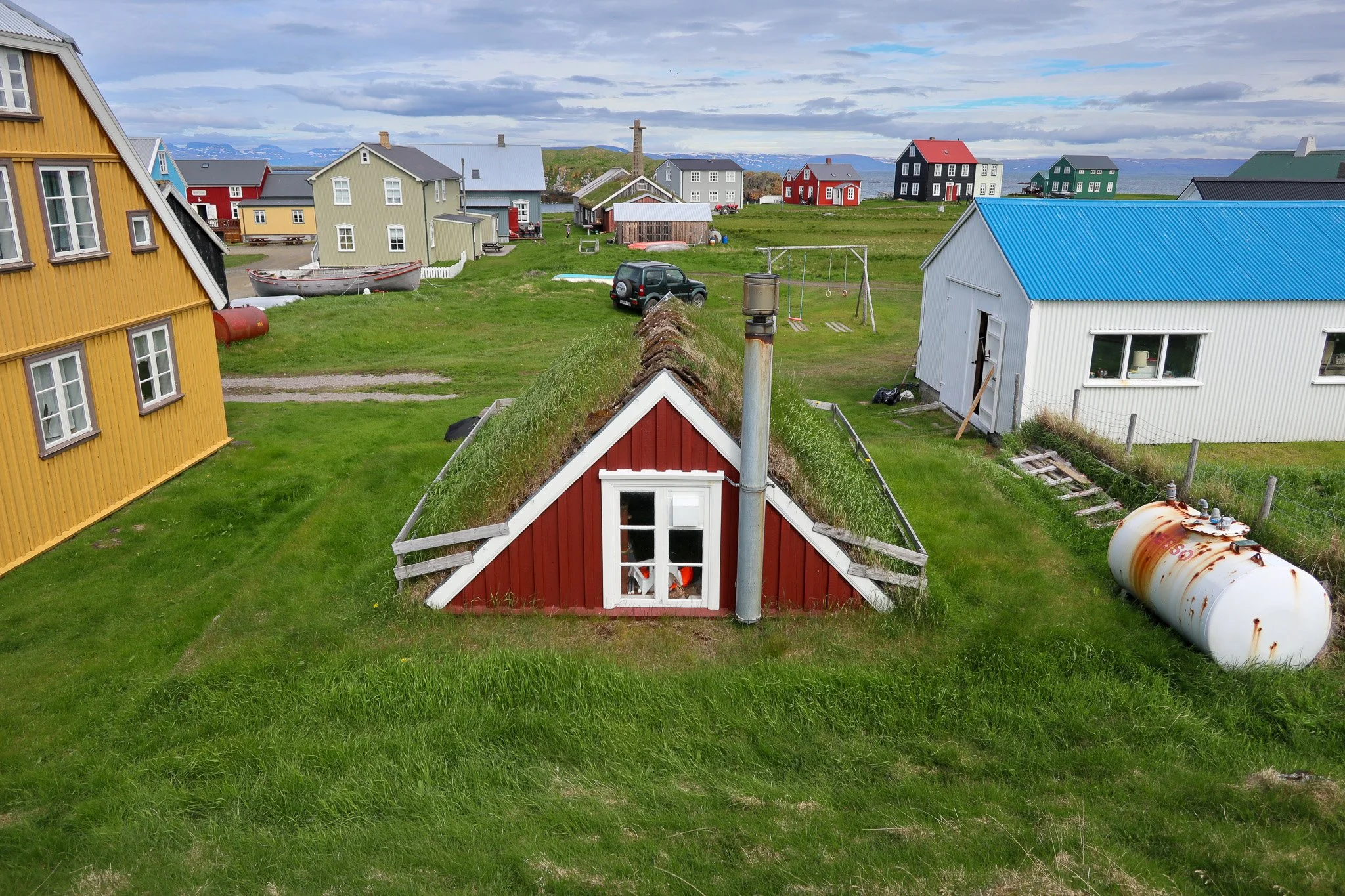 Colorful houses with grass roofs in a coastal village, with a white building having a blue roof and a rusty tank in the yard.