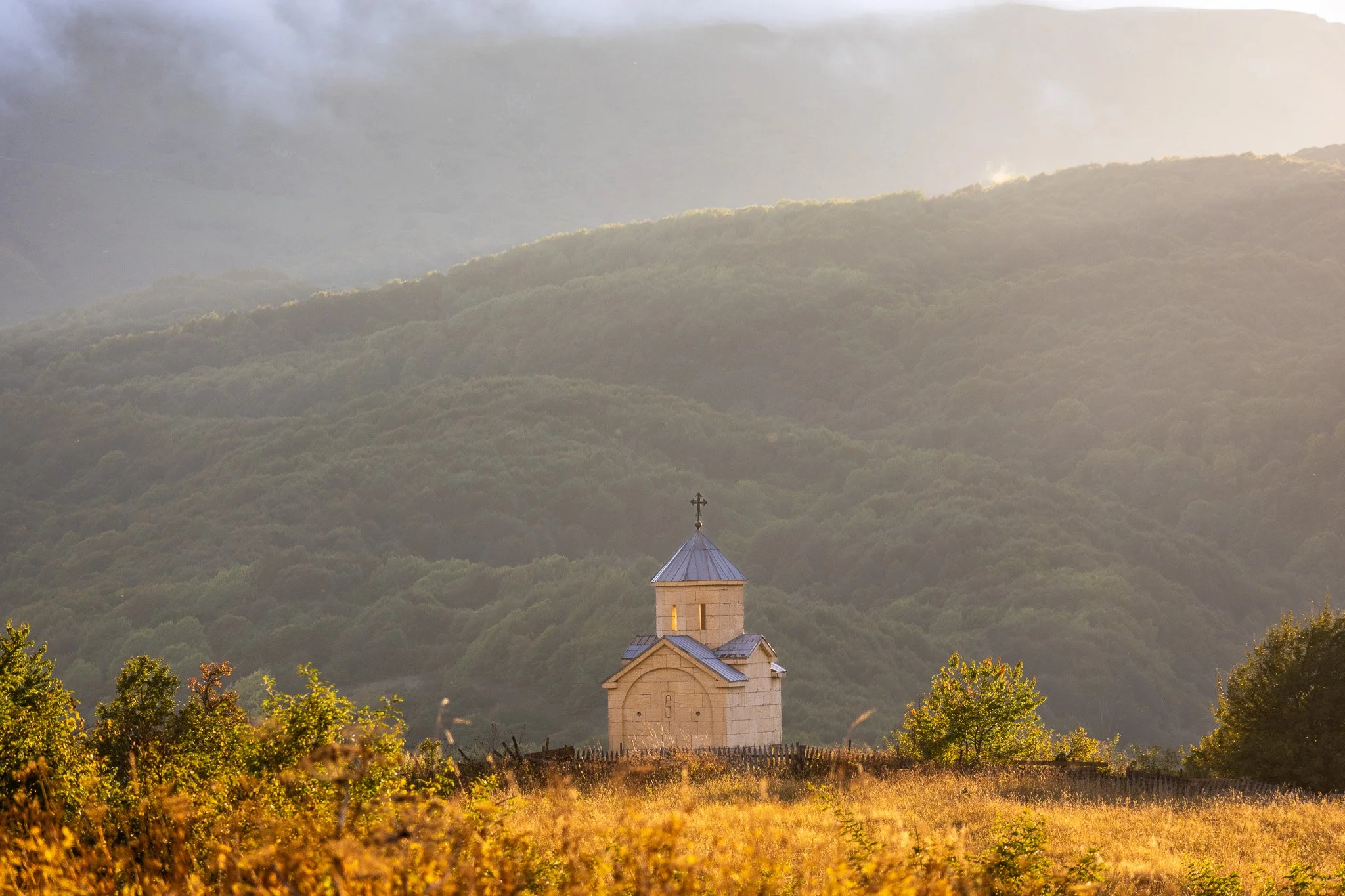 A small church with a cross on top, situated in a grassy field with trees, against a backdrop of green, rolling mountains under soft sunlight.