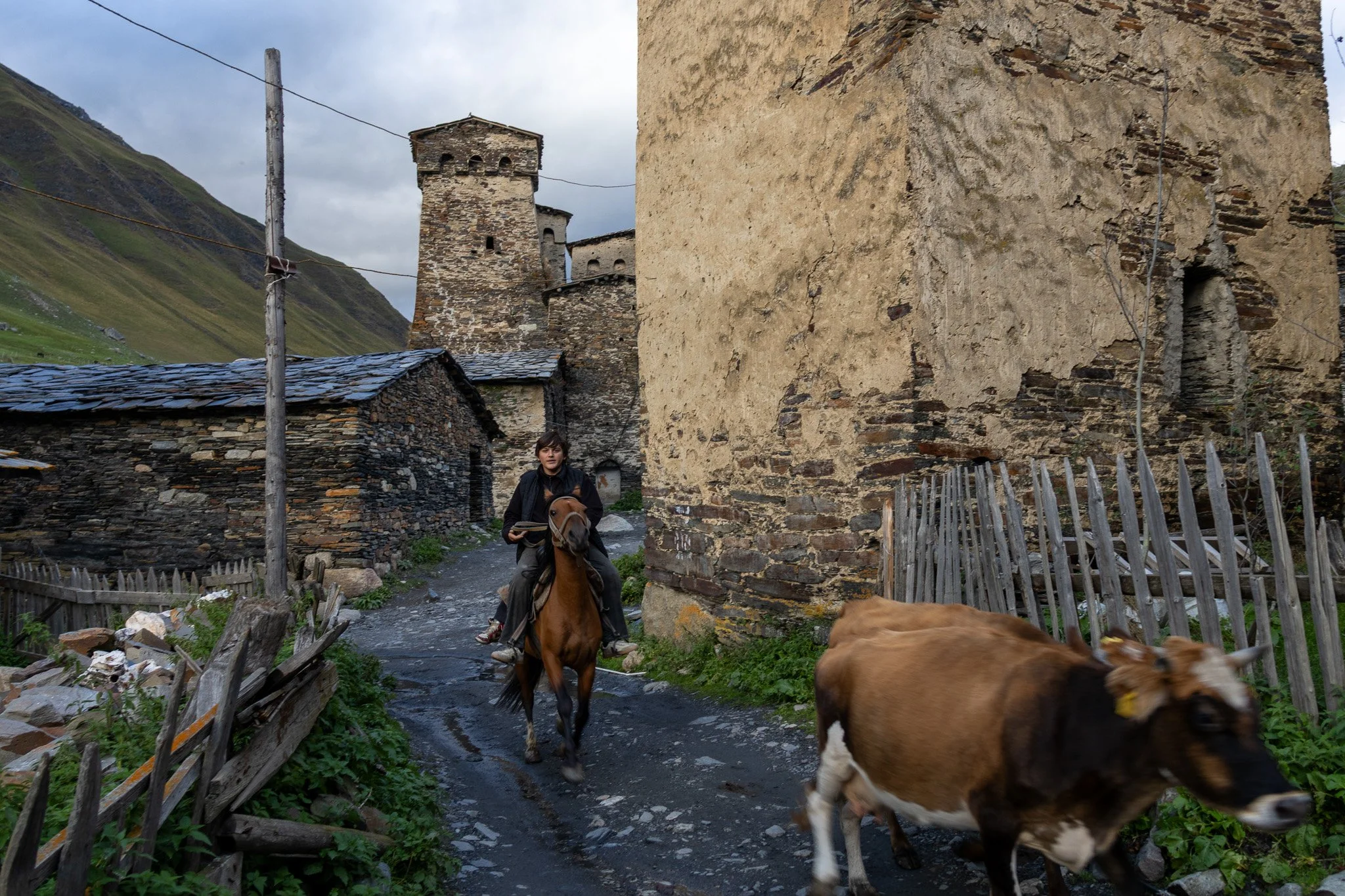 A person riding a horse on a narrow village street with old stone and wood buildings, a cow walking nearby, and green mountains in the background.