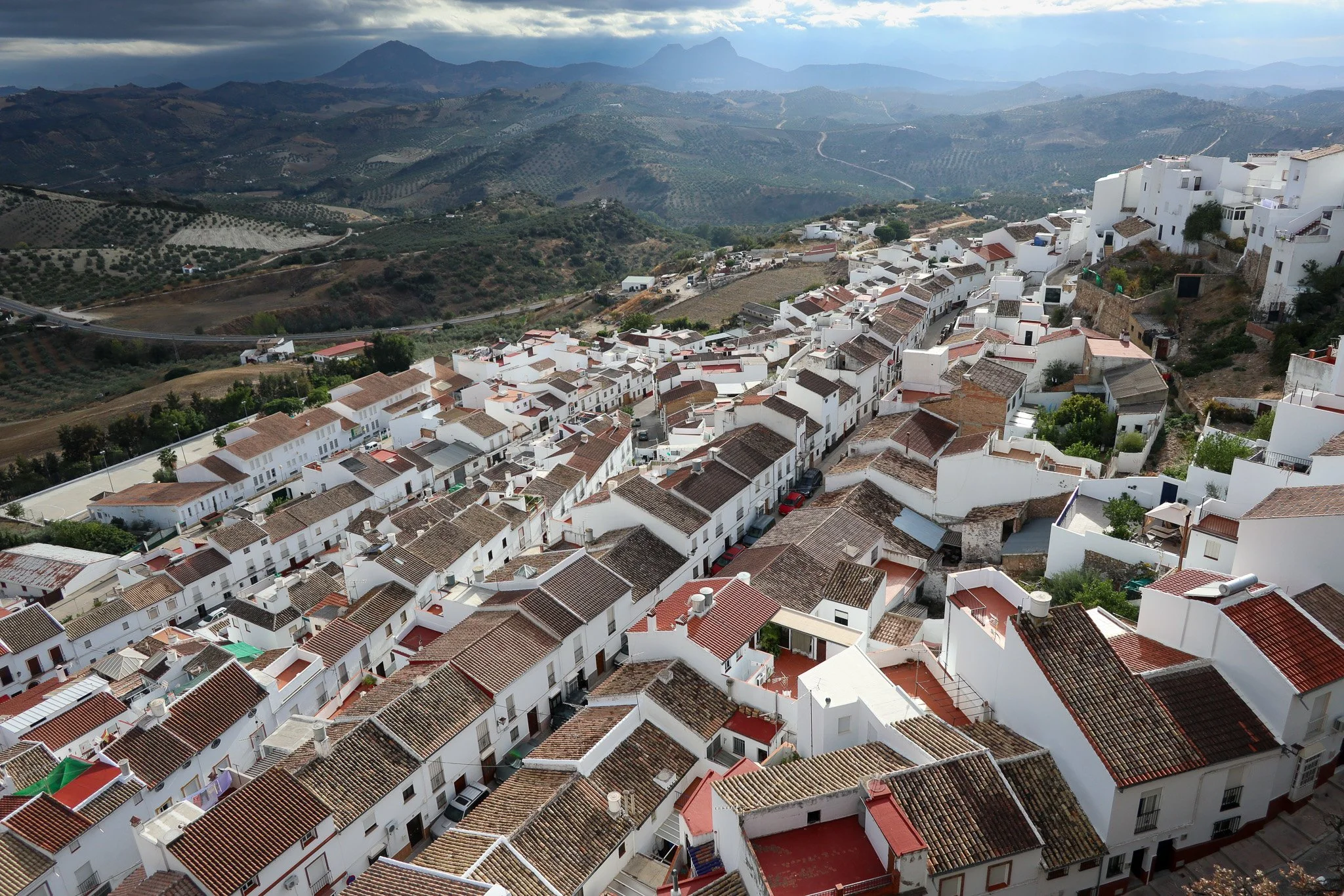 Aerial view of a hillside town with white buildings and reddish-brown tiled roofs, surrounded by green and brown hills, with mountains in the distance under a partly cloudy sky.