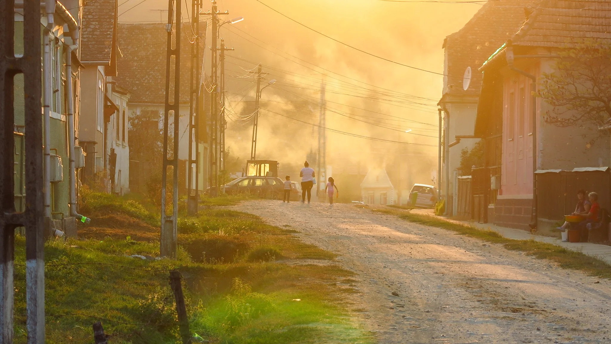 A rural street scene during sunset with a dirt road, houses on each side, utility poles with wires, a few children and adults walking, and two people sitting on a bench.