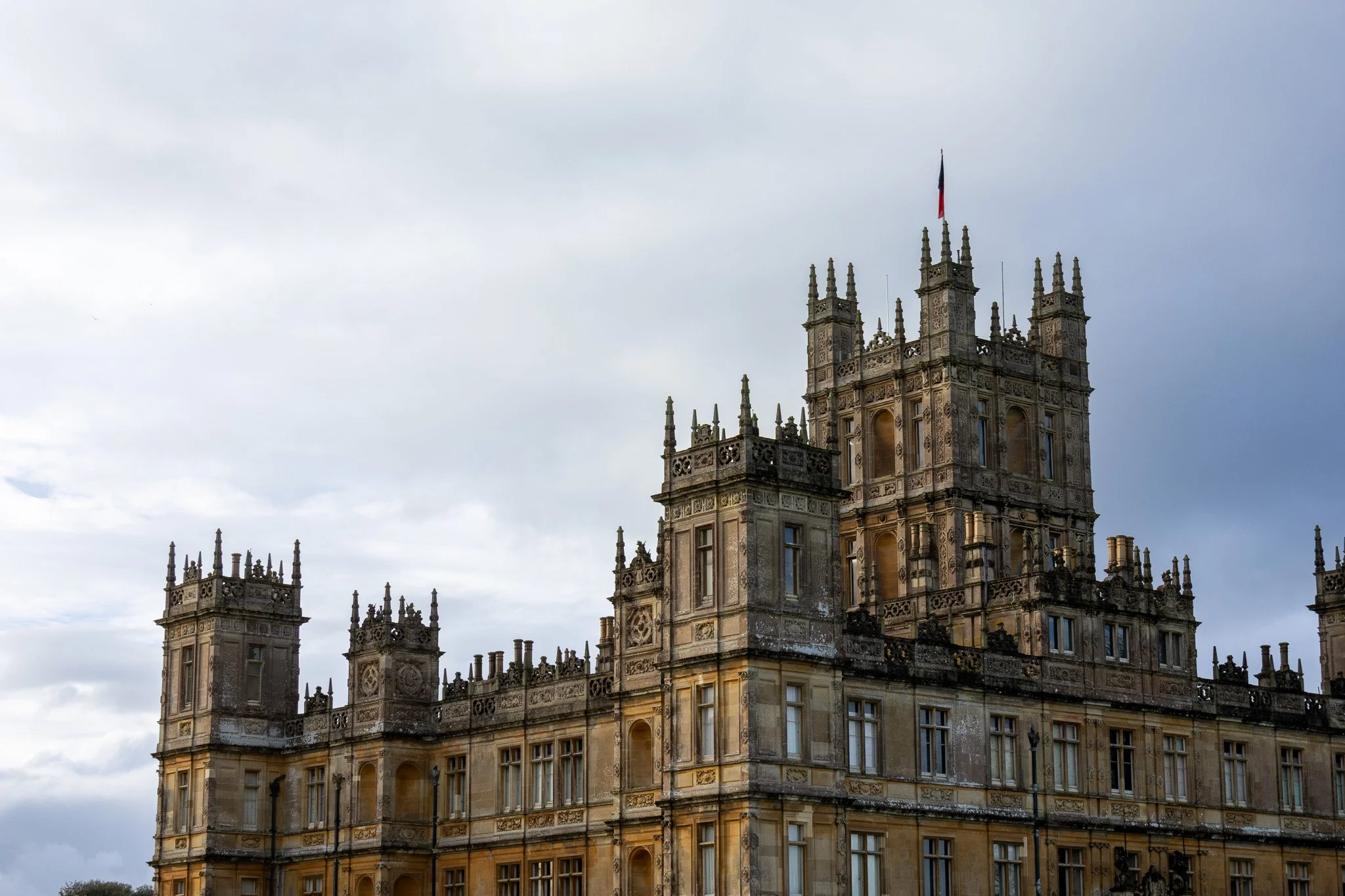 Close-up of a historic castle with ornate towers and pinnacles, featuring multiple windows and a flag flying atop the tallest tower, against a cloudy sky.