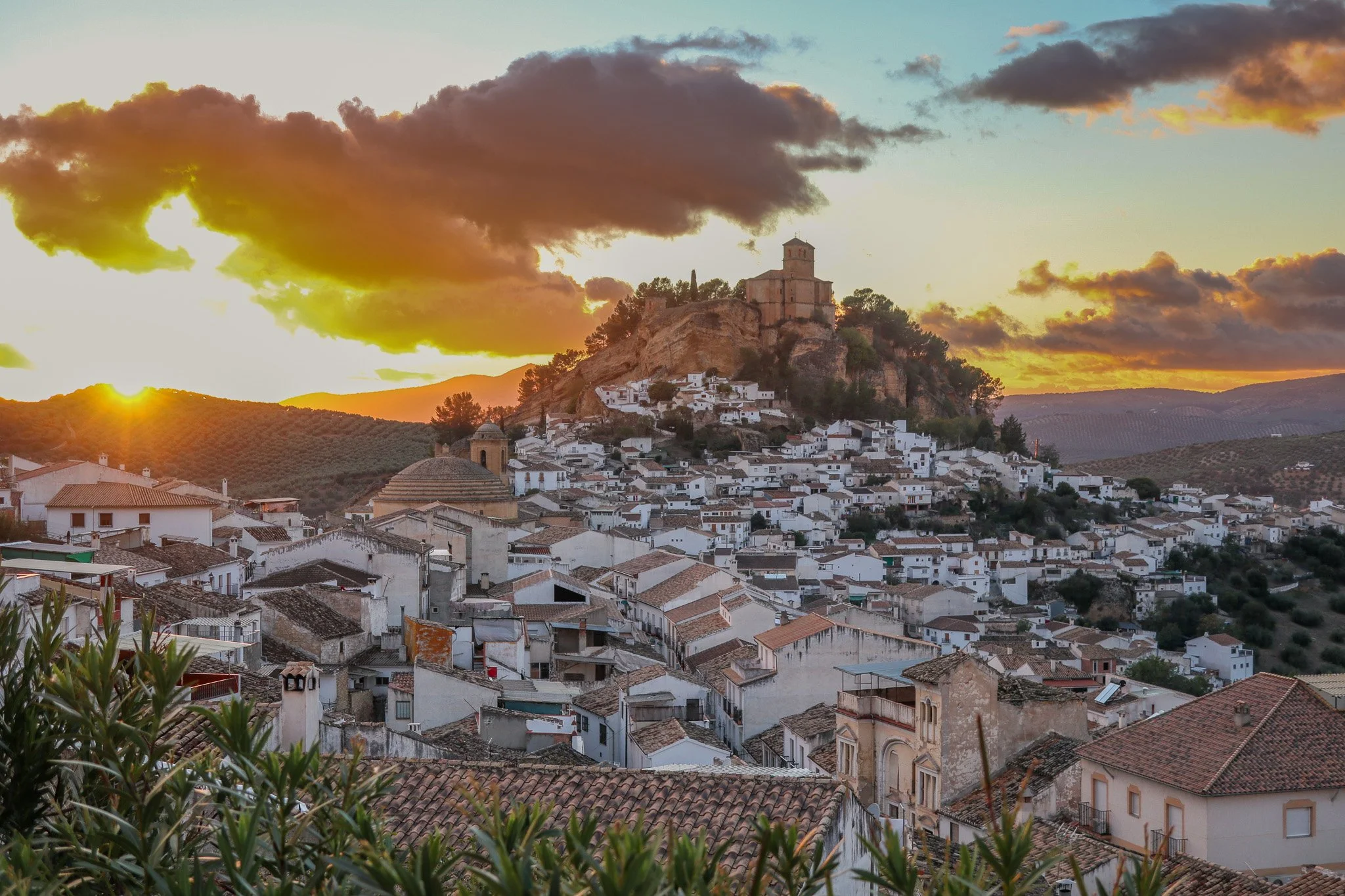 Sunset or sunrise over a hillside town with white buildings, a church, and a hilltop castle, with clouds in the sky.