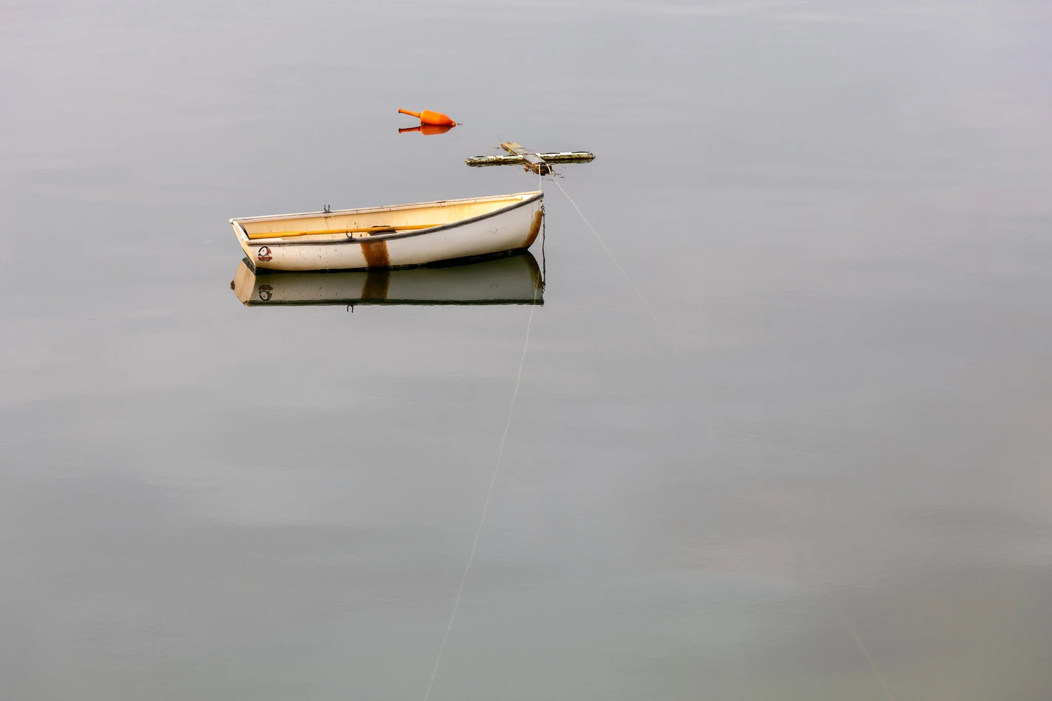 A small boat floating on calm water with a floating orange buoy nearby and a dock cleat. The water reflects the boat and buoy.