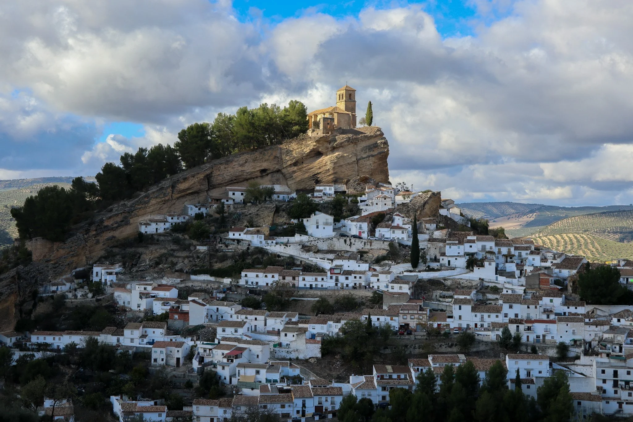 A hillside village with white houses and a church on top, surrounded by trees and rolling hills under a cloudy sky.