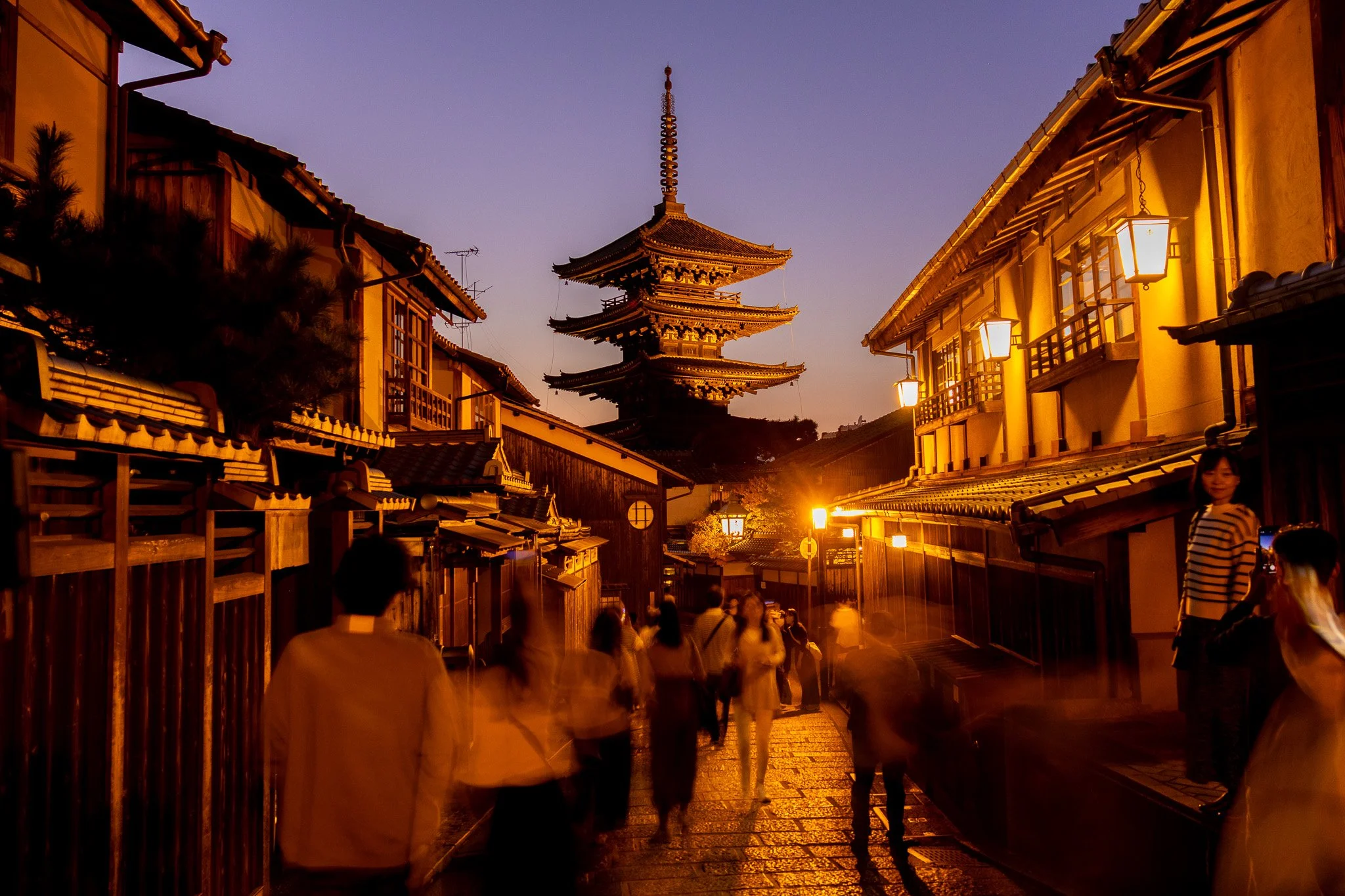 Night scene of a traditional Japanese street with wooden buildings illuminated by warm yellow lanterns and streetlights, featuring a multi-tiered pagoda in the background, with several people walking and taking photos.