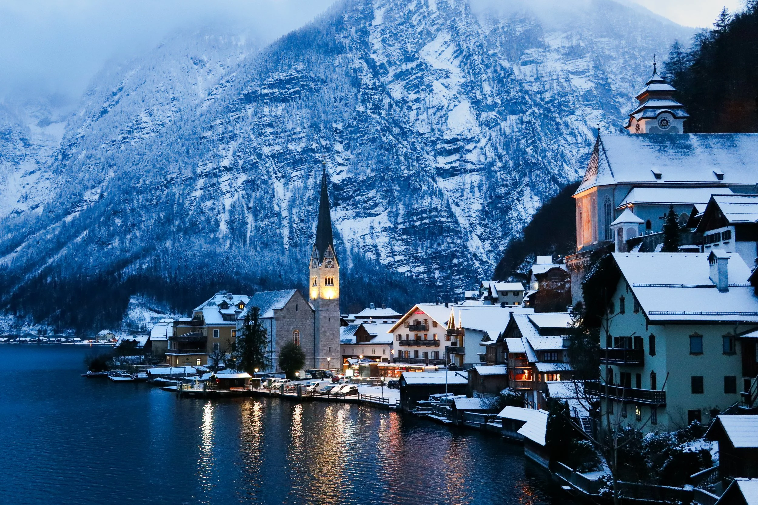 Snow-covered village by a lake with a church steeple and mountains in the background at dusk.