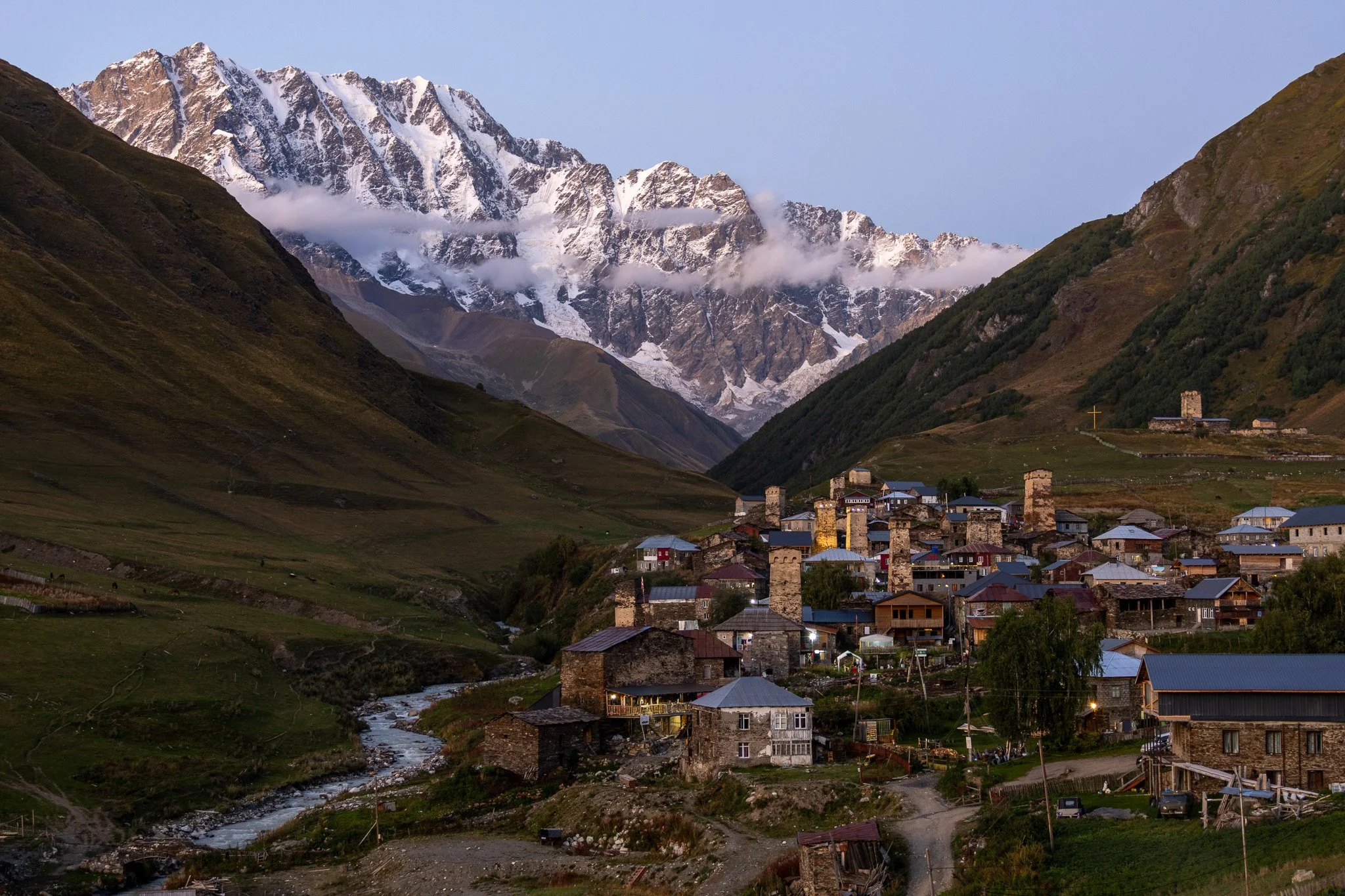 A mountain village at the base of snow-capped peaks, with a stream flowing through the valley and houses with stone walls and metal roofs.
