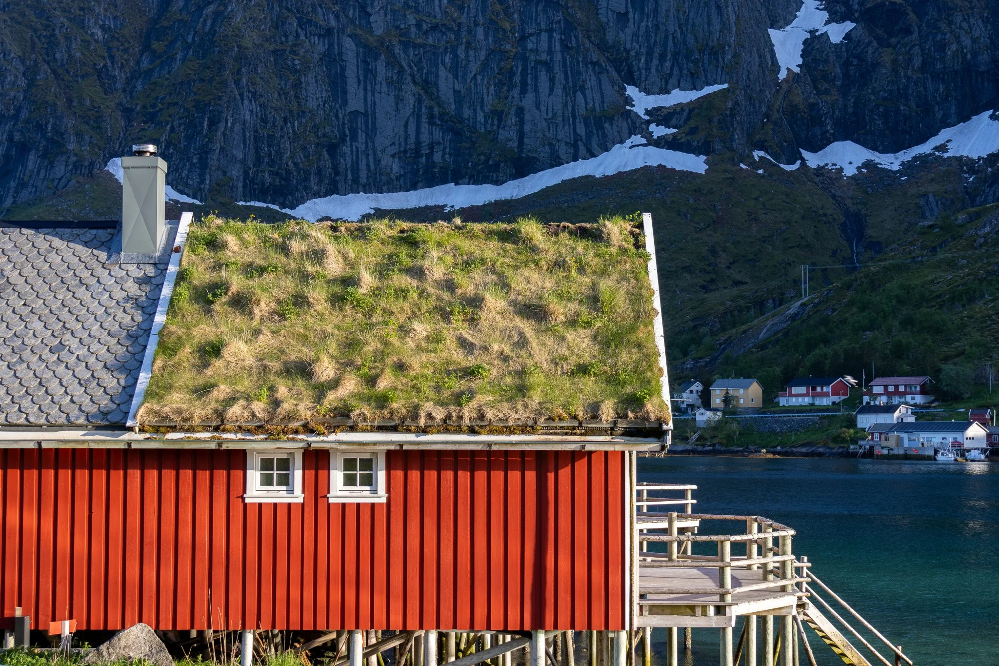 A red wooden house with a grassy green roof and two small white windows, located on stilts above the water, with a mountain and other houses in the background.
