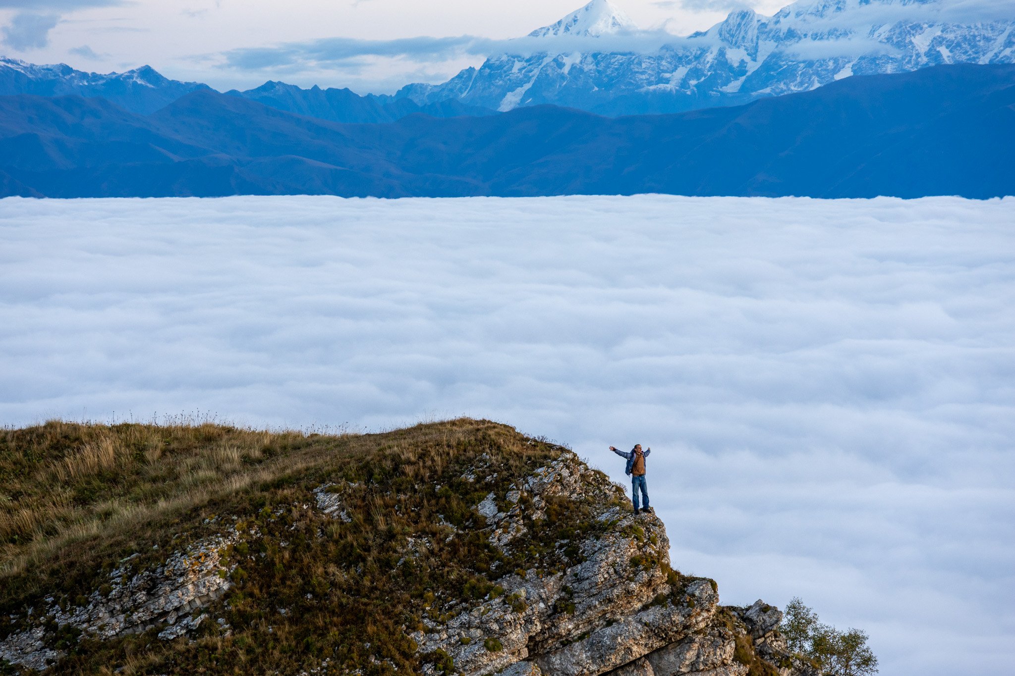 A person standing on a rocky mountain ridge with arms outstretched, overlooking a sea of clouds and distant snow-capped mountains.