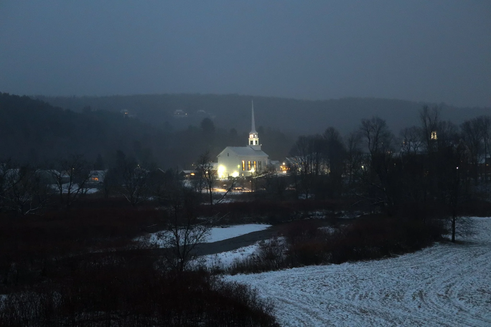 Nighttime view of a small town with a lit church and surrounding trees, snow-covered landscape, and distant hills in the background.