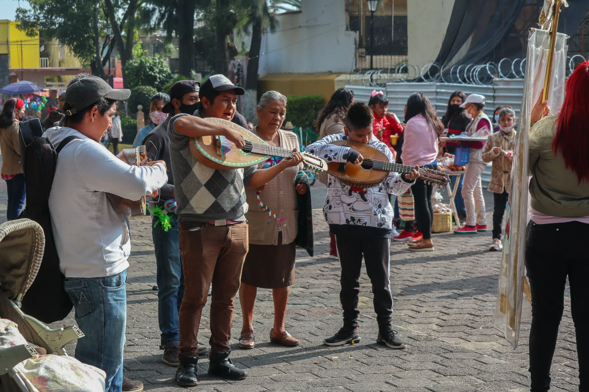 Group of people standing on a street playing guitars and singing, with others in the background selling items at a market.