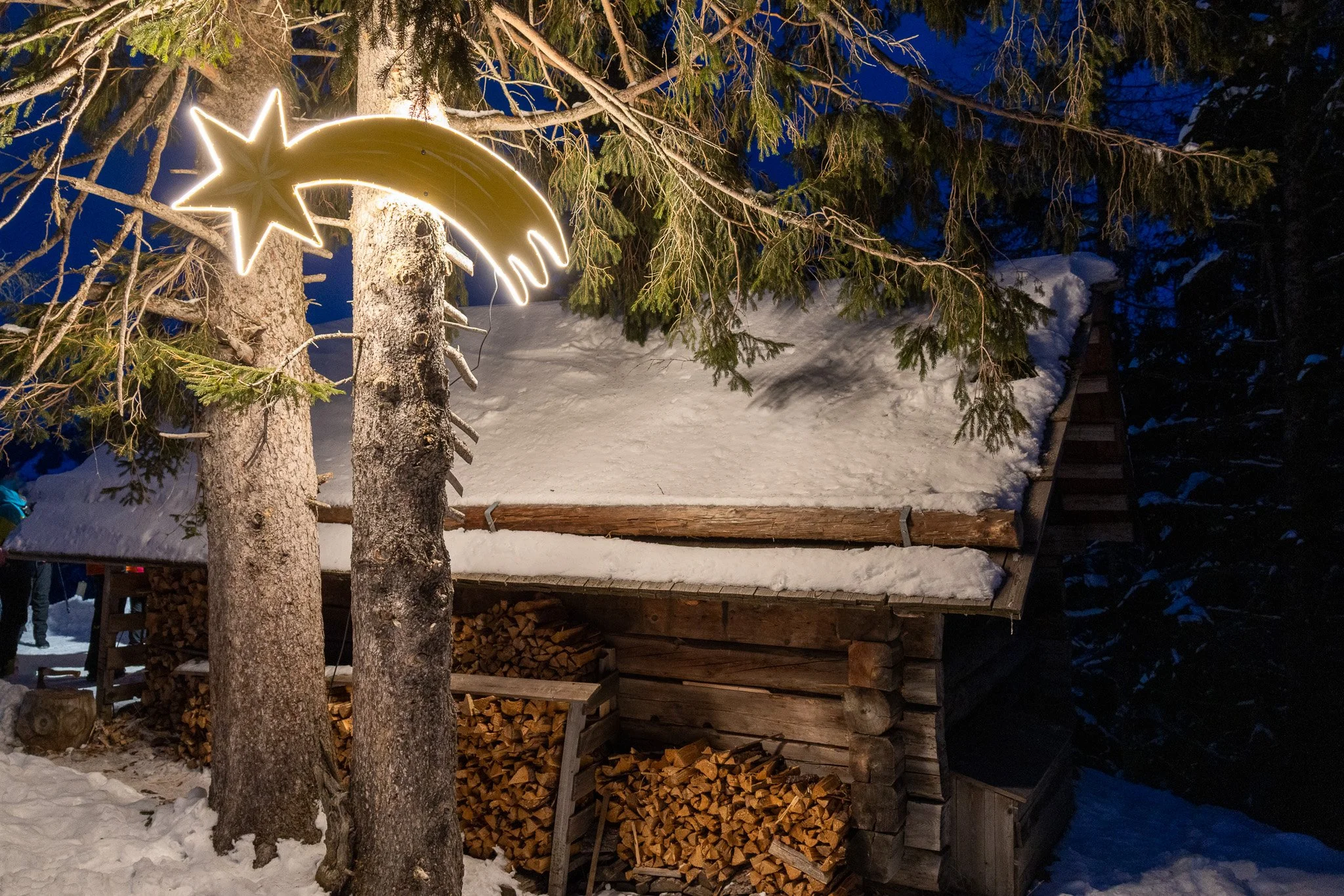 Snow-covered wooden cabin with stacked firewood underneath, illuminated star and shooting star holiday lights hanging from a tree branch.
