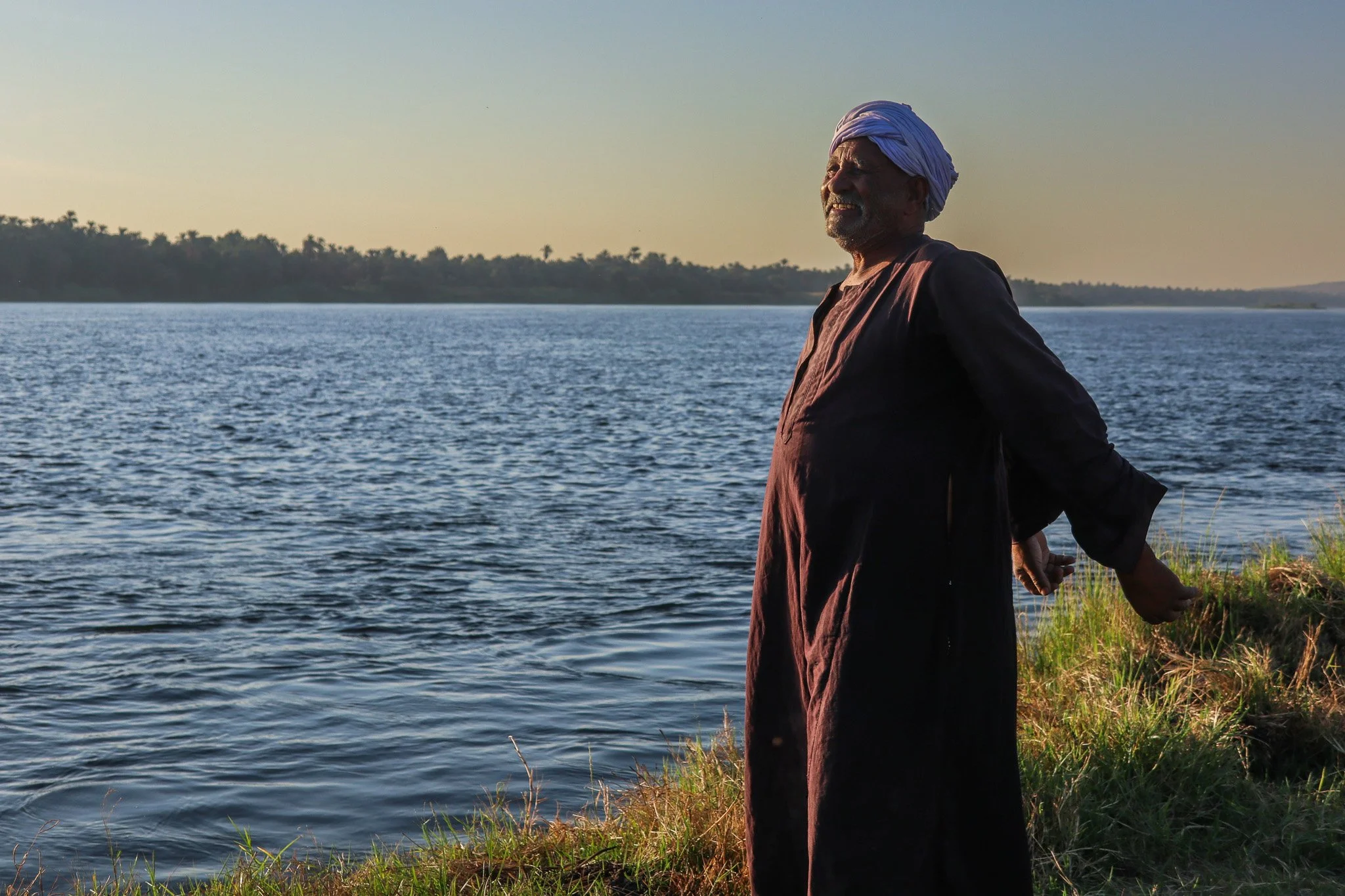 An elderly man with a white turban and traditional dark clothing stands by a river at sunset, gazing into the distance.