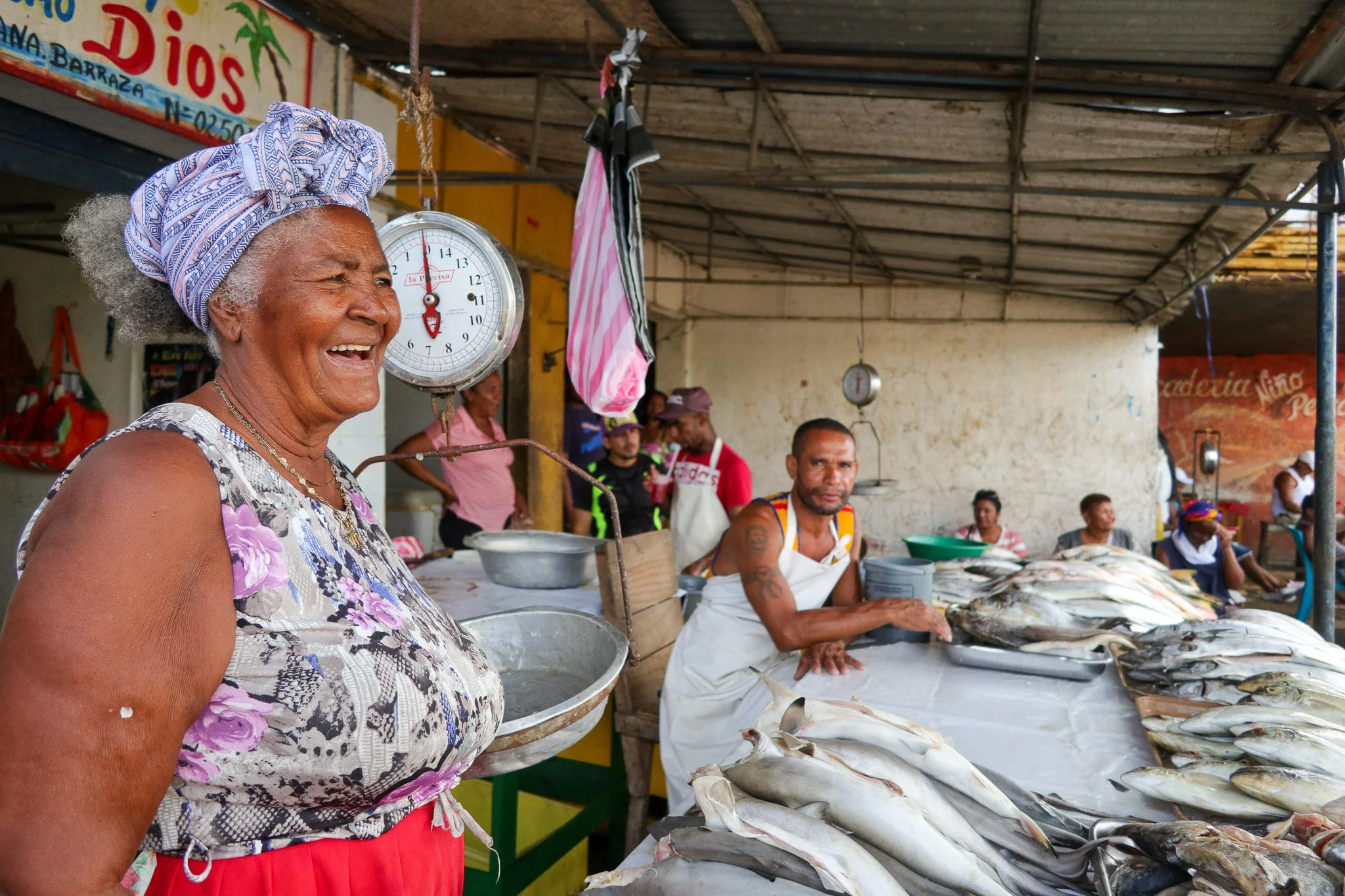 An elderly woman with gray hair, wearing a purple patterned headscarf and a sleeveless floral top, smiling at a fish market. Behind her are vendors and customers, with various types of fish displayed on a table.