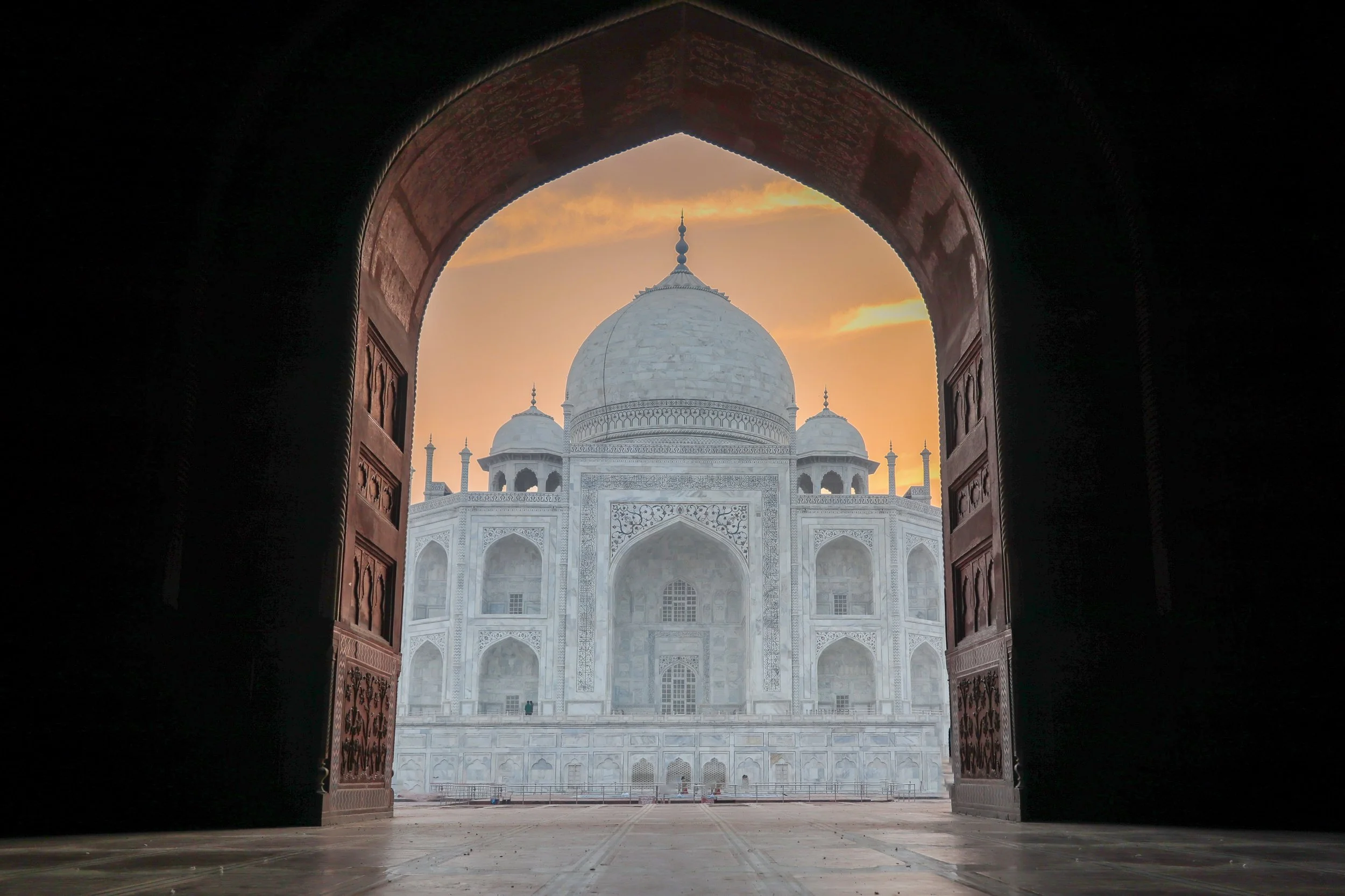 View of the Taj Mahal through an arched doorway at sunset, showcasing its white marble dome and minarets with a colorful sky in the background.