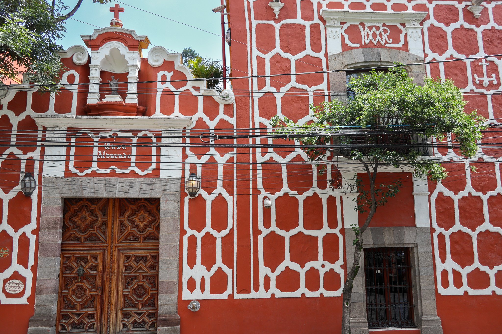 A decorative building with a red and white patterned facade, wooden door with intricate carvings, a small cross on top, a tree in front, and electrical wires crossing the scene.