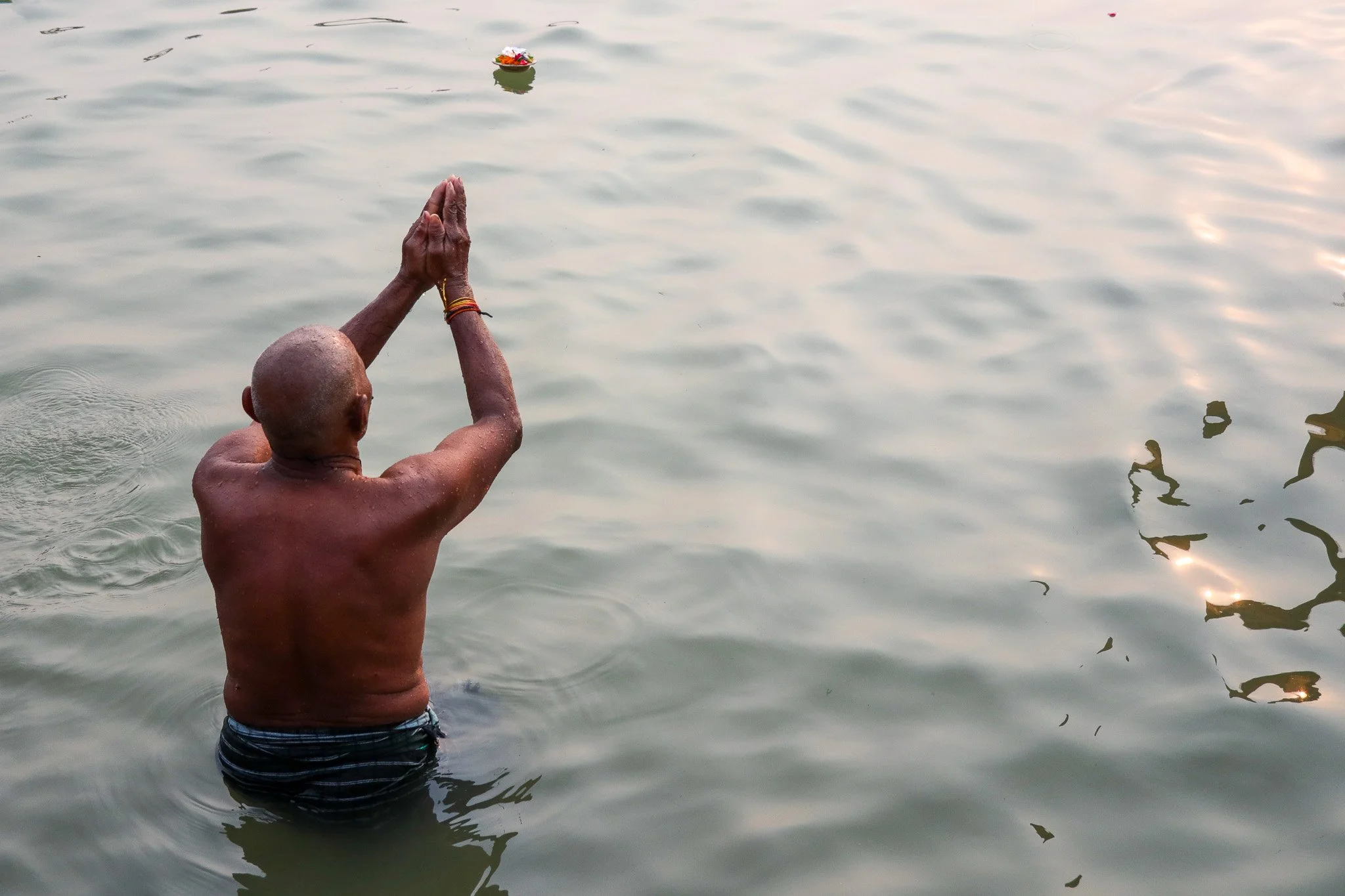 A man with a shaved head, standing waist-deep in water, performing a prayer with hands pressed together above his head. The water is calm, and the sky appears to be at sunset or sunrise, with reflections on the surface.