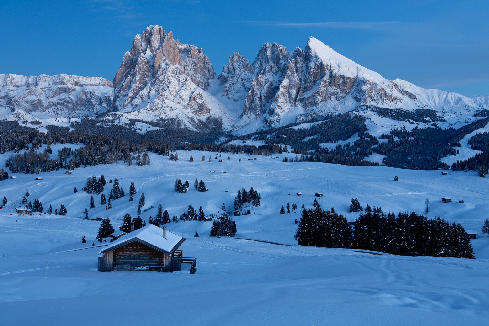 Snow-covered mountains with rocky peaks, evergreen trees scattered across rolling snow-covered hills, and a wooden cabin in the foreground during daytime.