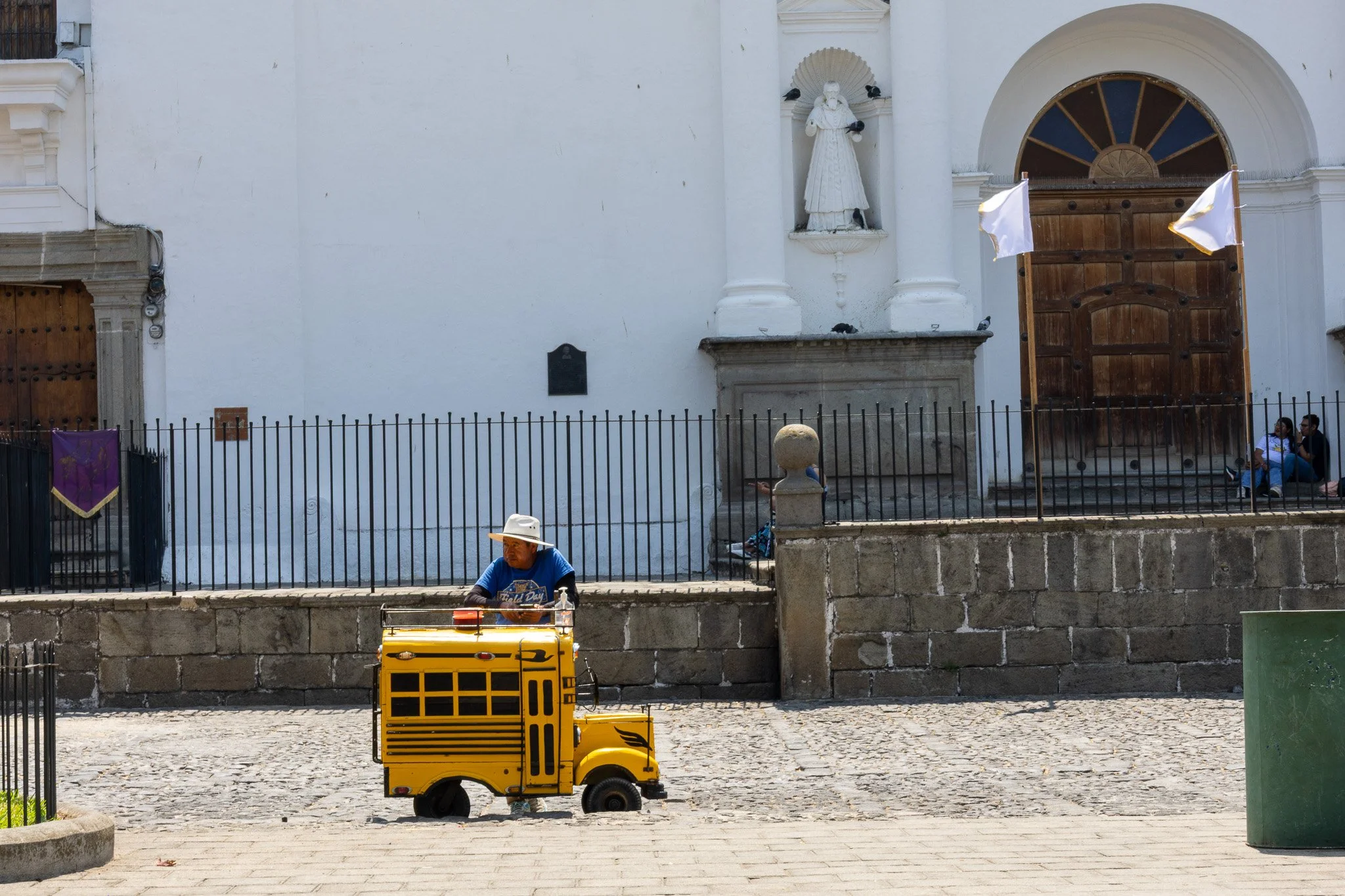 A street vendor in a blue shirt and white hat operating a yellow mobile food cart on a cobblestone sidewalk in front of a white church with a large wooden door, flags, and a statue in a niche.