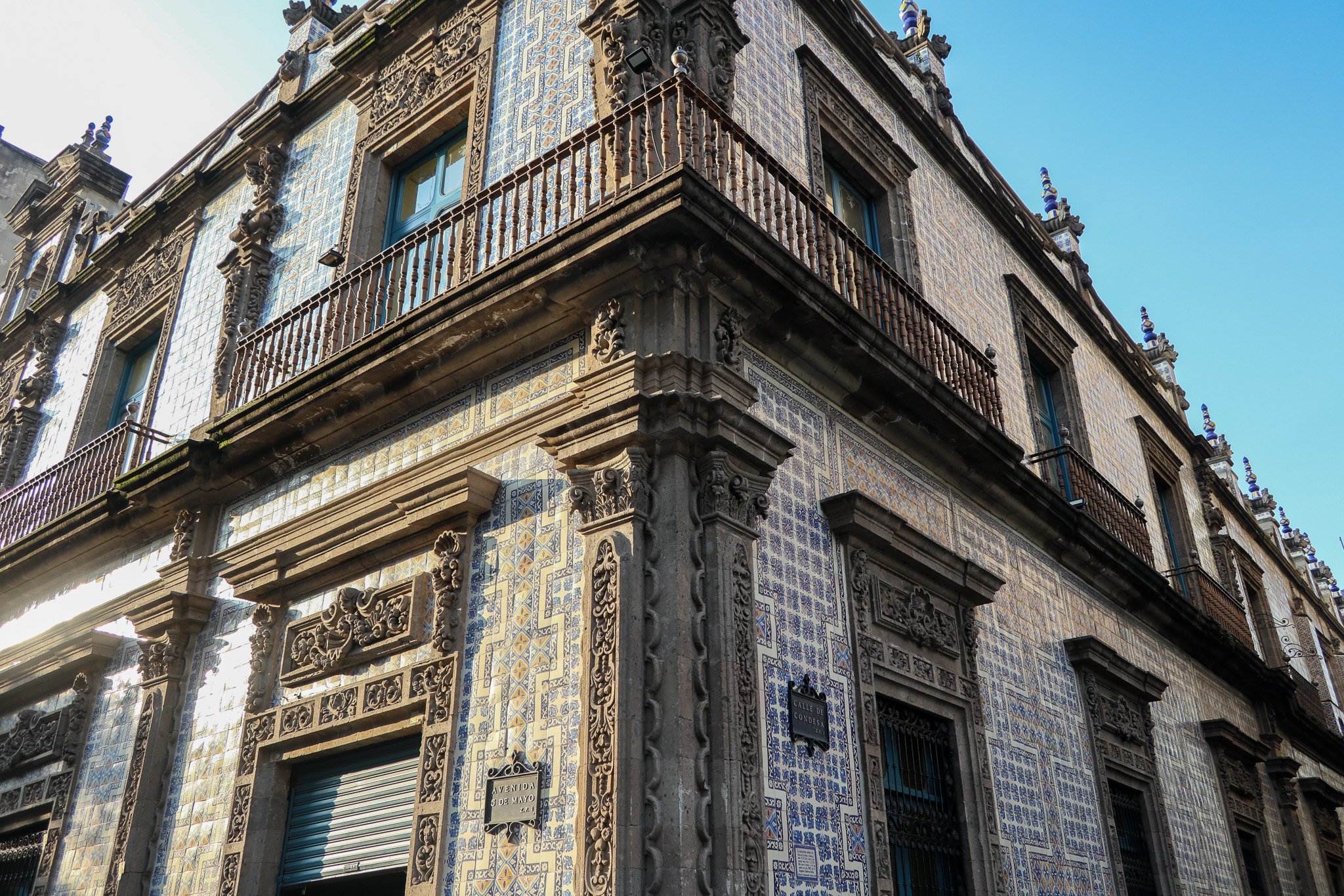 An ornate historic building with detailed stone carvings, blue and yellow decorative tiles, and wrought iron balcony railings, located on a corner at the intersection of Calle de Concordia and Avenida 5 de Mayo, with a bright blue sky above.