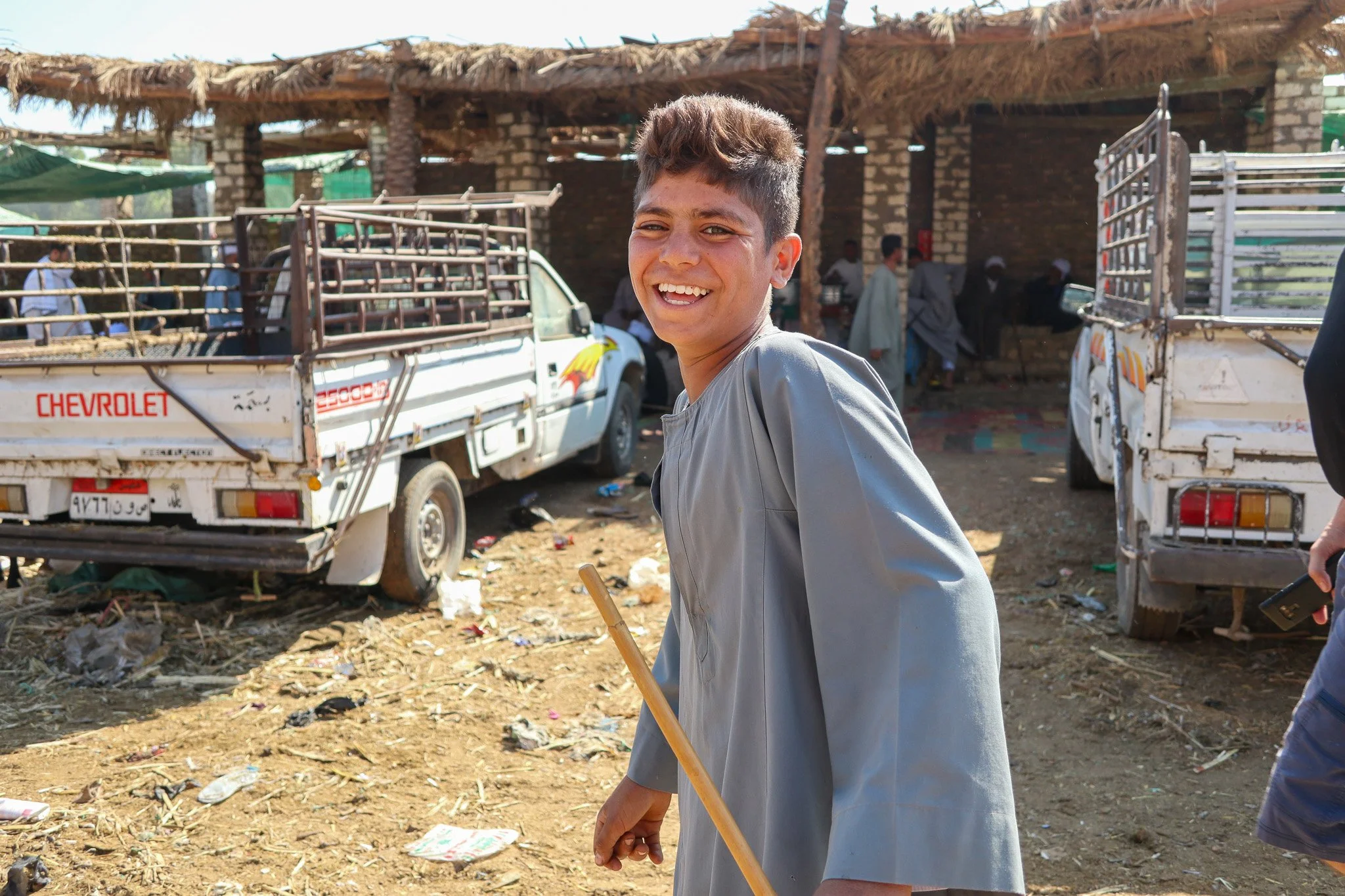 A young man smiling with a stick in hand standing outdoors in front of parked pickup trucks and a building with a thatched roof, in a dirt area with scattered trash, and several people in the background.