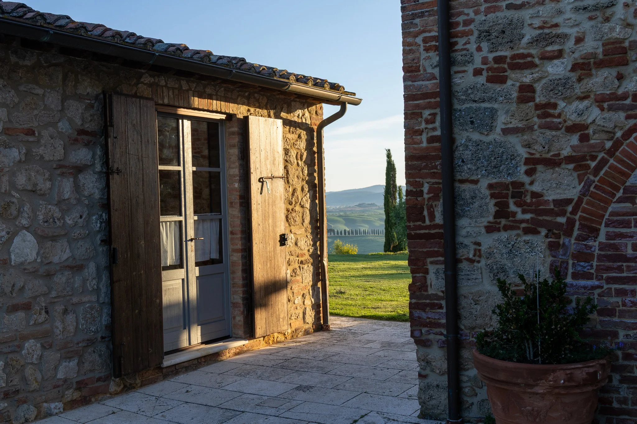 A stone and brick cottage door open to a view of the countryside with rolling hills, trees, and a blue sky.