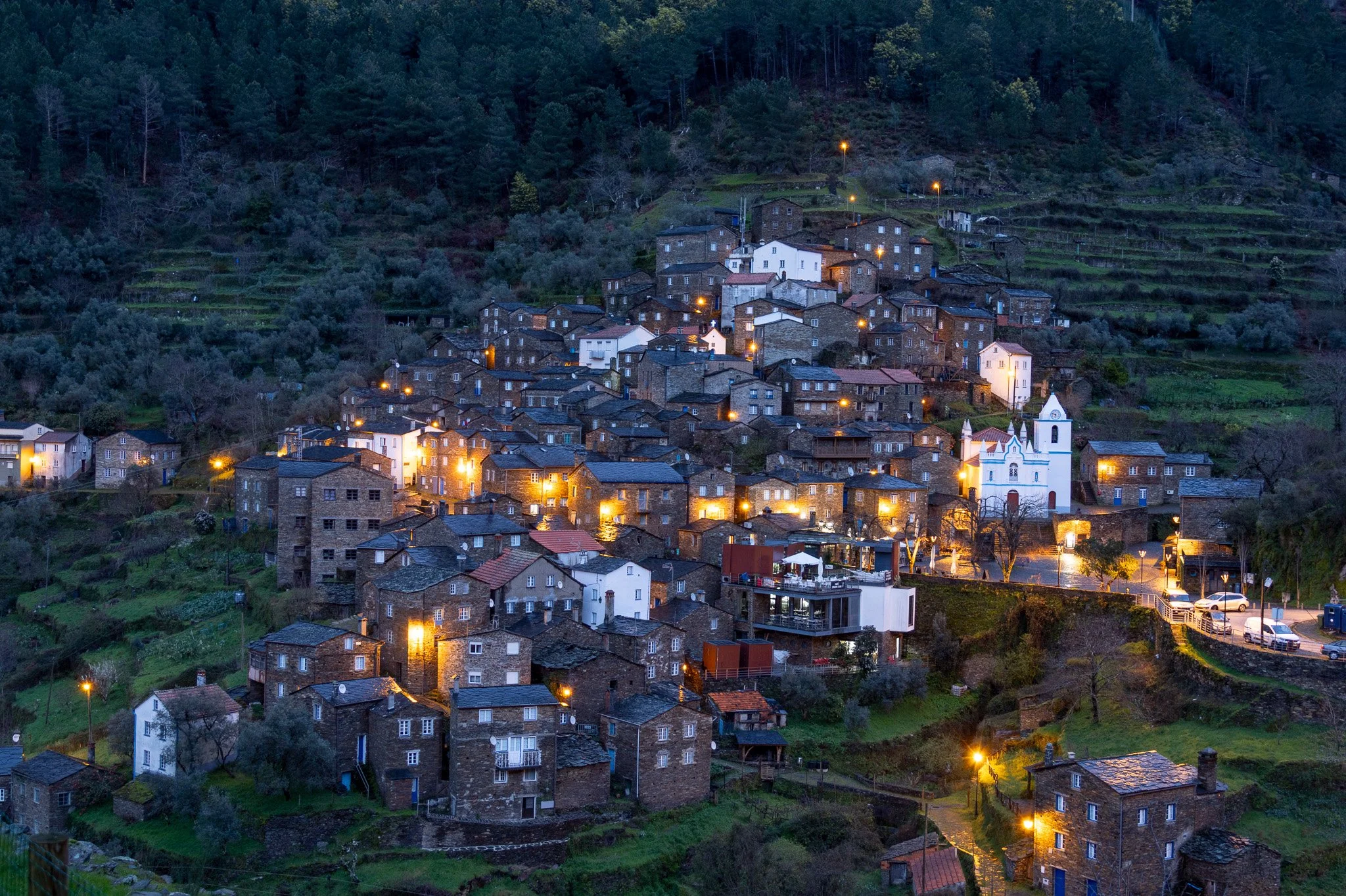Nighttime view of a hillside village with closely packed stone houses, some with lights turned on, surrounding a white church with a bell tower, located in a green forested area.
