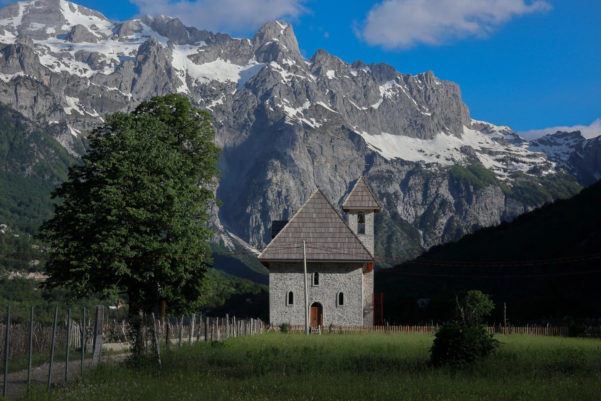 A stone church with a wooden roof and bell tower sits in a green field, with large snowy mountains in the background under a blue sky.