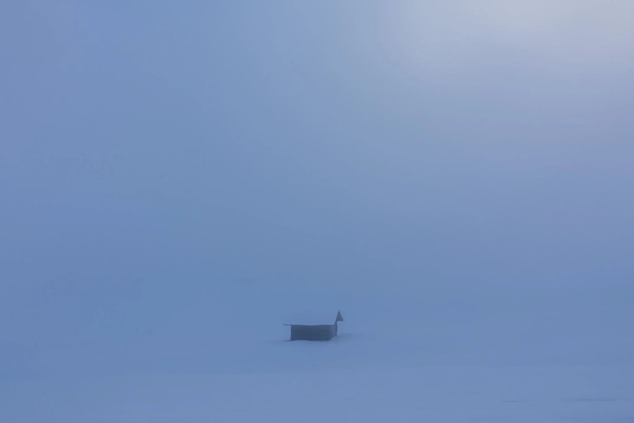 A small, wooden boat floating alone on a foggy, snow-covered body of water or frozen landscape, with minimal visibility and no other visible objects.
